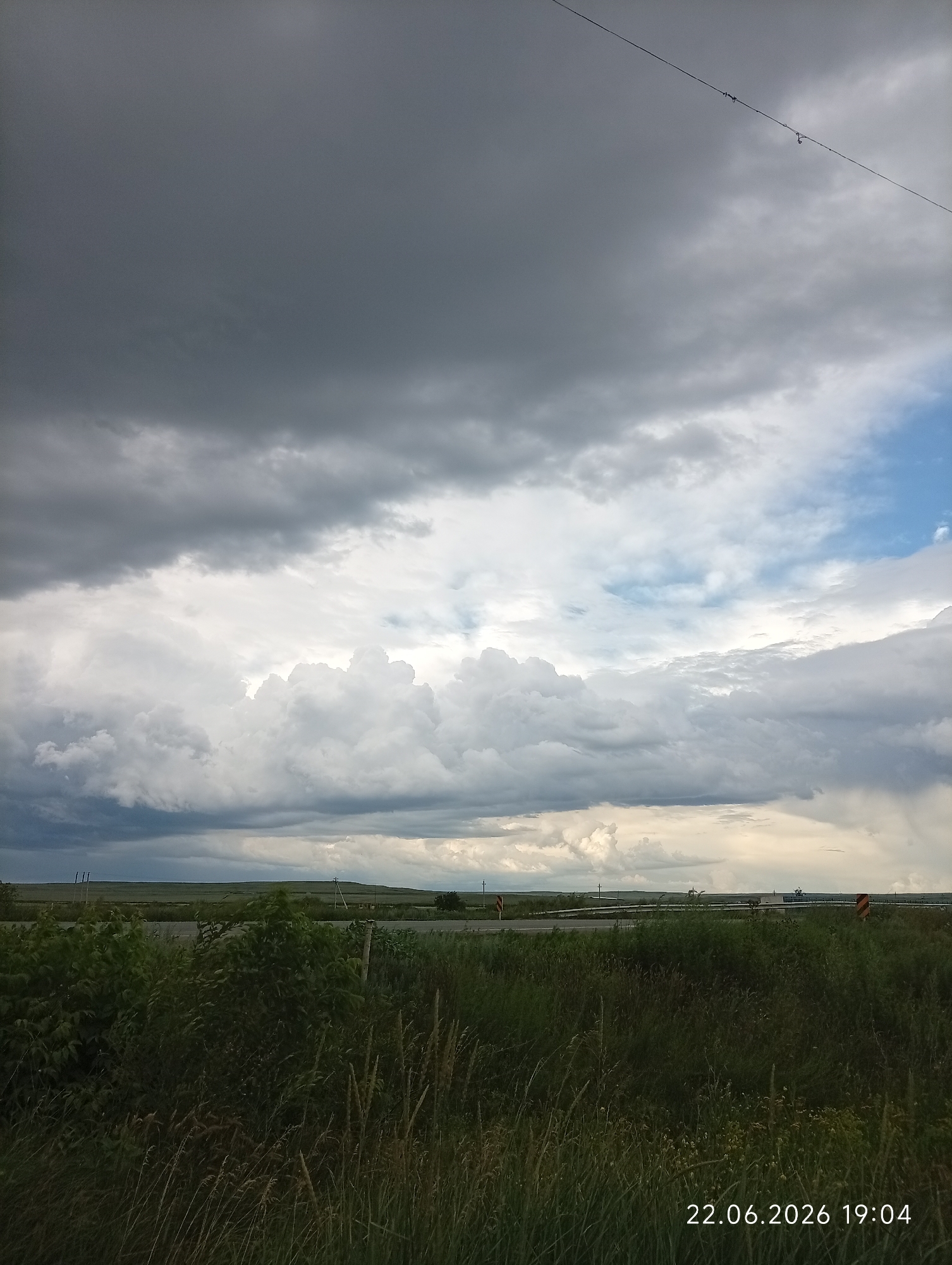 The image depicts a cloudy sky over a rural landscape, likely on the outskirts of Sarytogay, Kazakhstan. The sky is dominated by dramatic storm clouds, with dark gray formations in the upper portion and lighter, more voluminous clouds lower down. Patches of blue sky are visible in the upper right quadrant. A thin wire runs diagonally across the upper part of the frame.
Below the sky, a flat, grassy plain stretches to the horizon. A road, possibly a highway, is visible in the mid-ground, with some traffic infrastructure like signs and barriers. In the foreground, there is dense, tall grass and foliage. The overall impression is one of an impending storm or recent rainfall in a natural, undeveloped area. The timestamp "22.06.2026 19:04" is overlaid on the bottom right corner of the image. There are no people or animals visible.