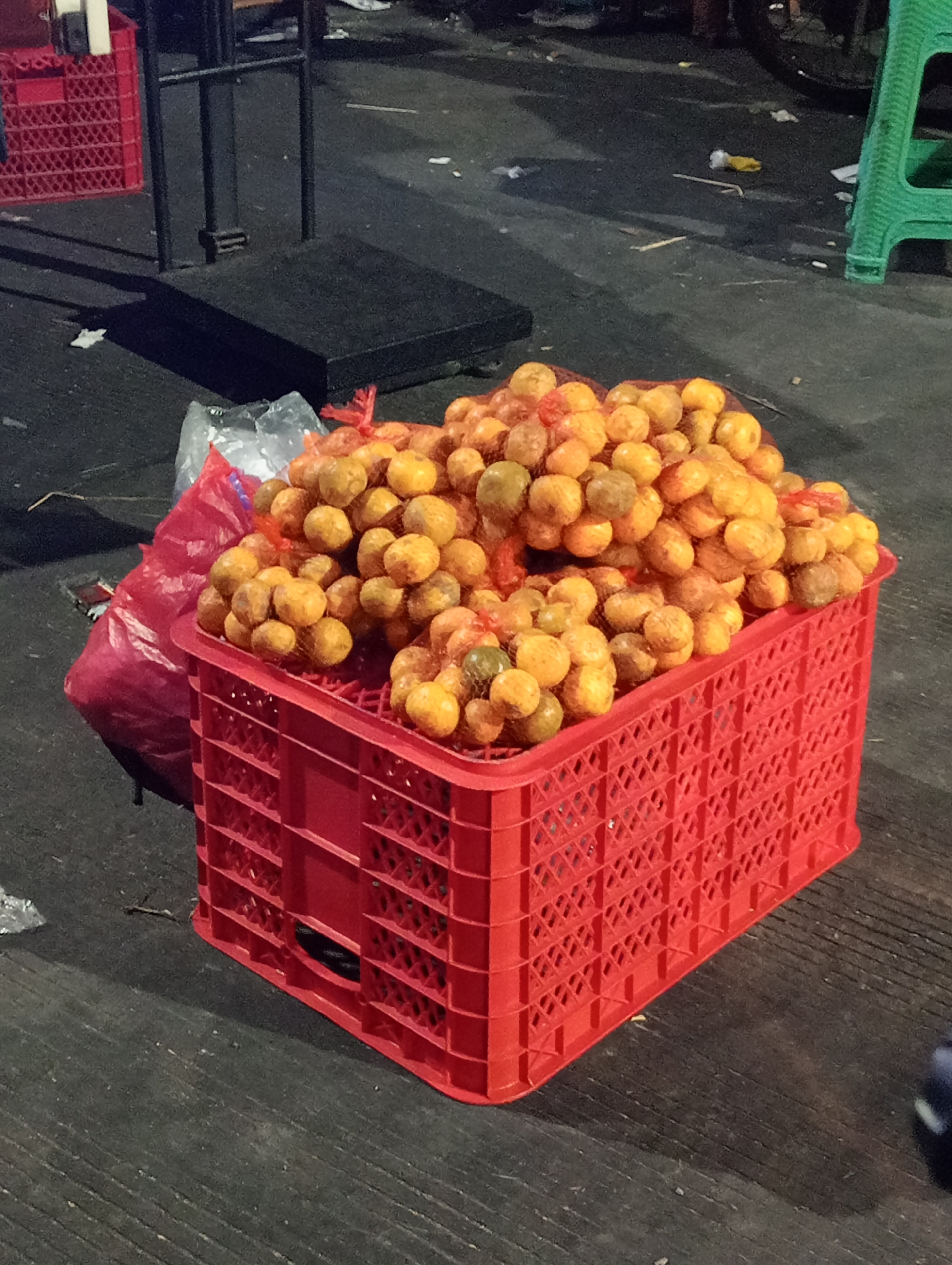 A close-up, eye-level, outdoor shot shows a red plastic crate filled with small, netted bags of oranges. The crate is on a dark, paved surface, possibly a street or market area. In the background, a black scale or platform is visible, along with red and green plastic crates. The scene suggests a market setting, possibly in the city of Demak, Indonesia, given the oranges are a common fruit sold in such locations. The lighting suggests it might be evening or a dimly lit area. No people are visible in the frame.