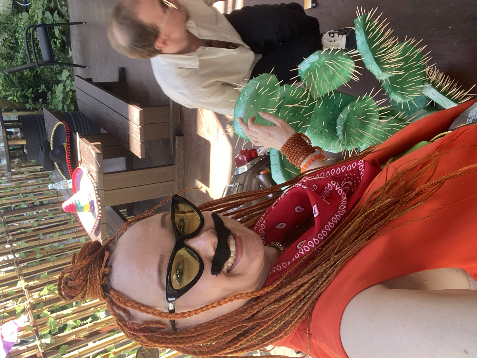 The scene depicts an outdoor gathering in Sochi, Russia, with two individuals and thematic decorations. In the foreground, a light-skinned woman with long, orange-red braided hair, styled partly in a high bun, directly faces the camera, capturing a selfie. She wears black cat-eye sunglasses with yellow-tinted lenses, a fake black mustache, a bright orange sleeveless garment, and a red bandana with white paisley patterns around her neck. Her left wrist is adorned with multiple amber and orange beaded bracelets, and her left hand is extended towards a large, green artificial cactus prop with numerous light-colored spikes, positioned on the ground. Behind the woman and to her right, a light-skinned man with short, light brown hair and clear-rimmed glasses is observed in a crouching posture, looking down and to his right, wearing a light-colored collared shirt, dark pants, and black and white sneakers. The background features a wooden picnic table with attached benches, on which a colorful sombrero, a black sombrero, and a plastic water bottle are placed. A bamboo-style fence with climbing green foliage, additional greenery, and a black metal chair are visible further in the background. The ground surface is dark and appears to be paved or concrete, under bright daylight.