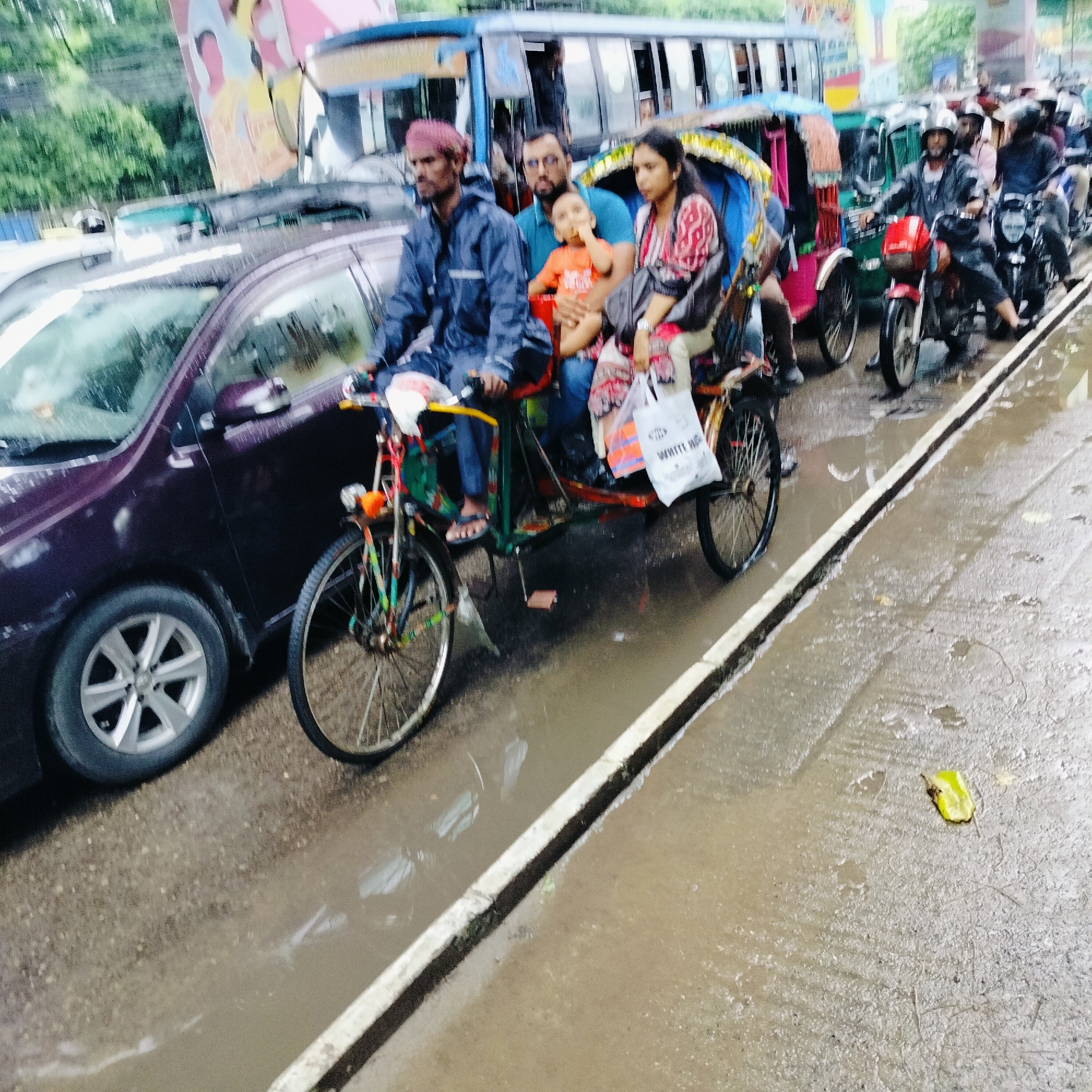 The image depicts a street scene in Dhaka, Bangladesh, likely during or after a rainfall, as evidenced by the wet pavement and reflections on the road surface. A purple car is parked on the left side of the frame, its side mirror visible. In the center, a cycle rickshaw is prominent, carrying a driver and passengers. The driver, wearing a dark blue raincoat and a pink turban, is pedaling the rickshaw. Seated behind him is a man with glasses, holding a young child in an orange shirt. A woman in a patterned shirt is seated next to the man. A white shopping bag with text is visible in the rickshaw. Various other vehicles, including a bus and motorcycles, are present in the background, suggesting traffic congestion. The environment appears to be an urban setting with trees visible in the upper left corner.