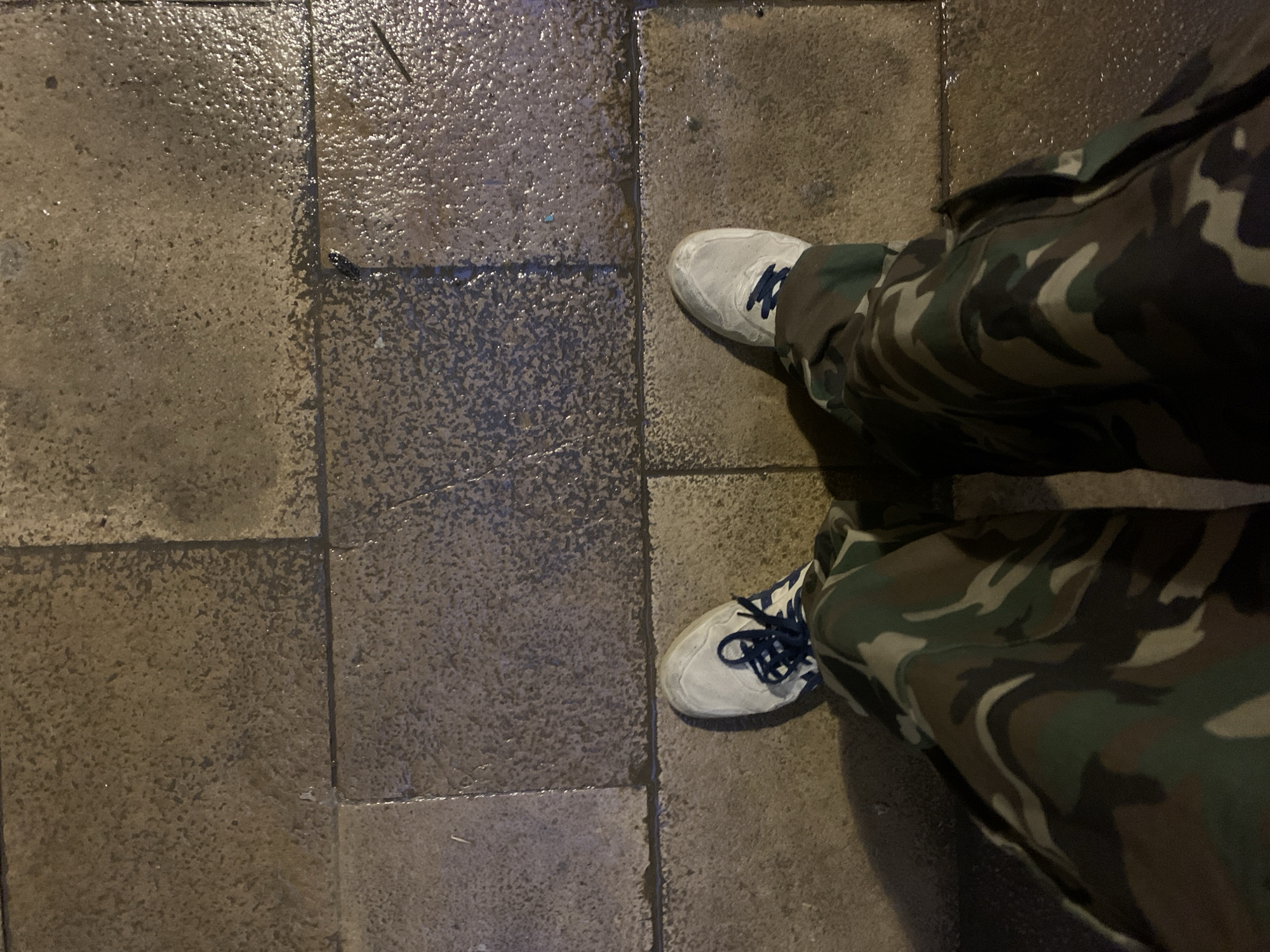 The image shows a person standing on a paved sidewalk, likely in an urban area. The sidewalk is composed of gray concrete tiles, some of which appear wet, suggesting it may have rained recently. The person is wearing camouflage pants and white sneakers with blue laces. The scene is dimly lit, likely at night, as evidenced by the shadows and wet appearance of the tiles. Based on the type of floor texture in the foreground, it is not possible to infer anything concrete about the location of Barcelona.