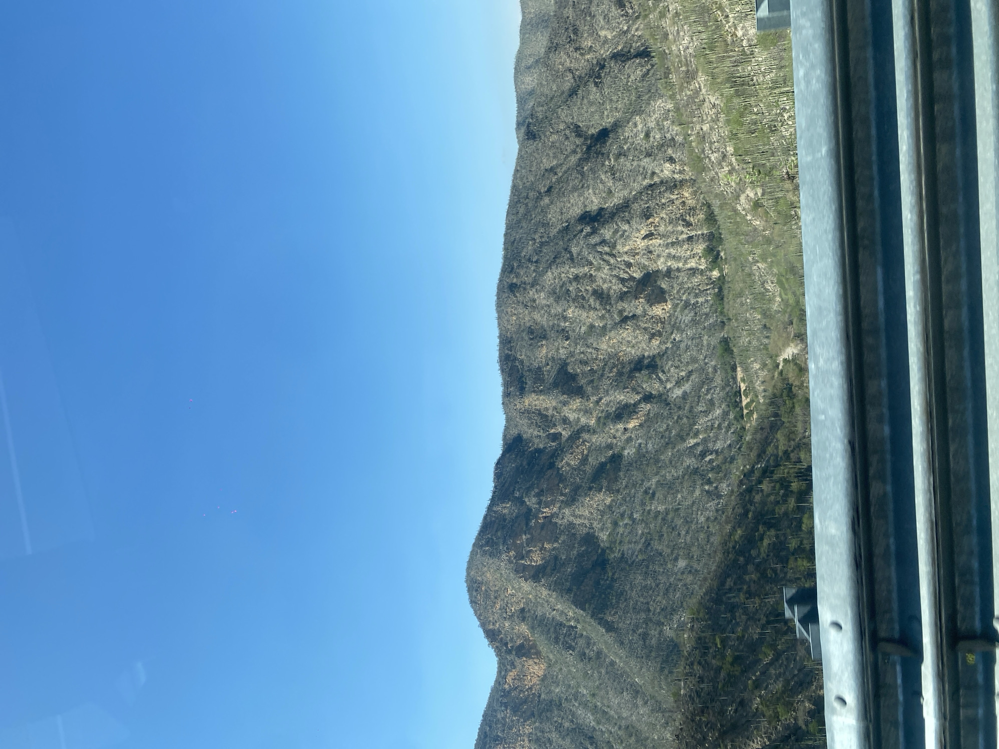 The image shows a mountain in Cerro Verde, Mexico, under a clear blue sky. The mountain has sparse vegetation, possibly cacti and shrubs, covering its rocky terrain. In the foreground, part of a metallic structure with vertical bars and what looks like supports is visible on the right side of the image.
