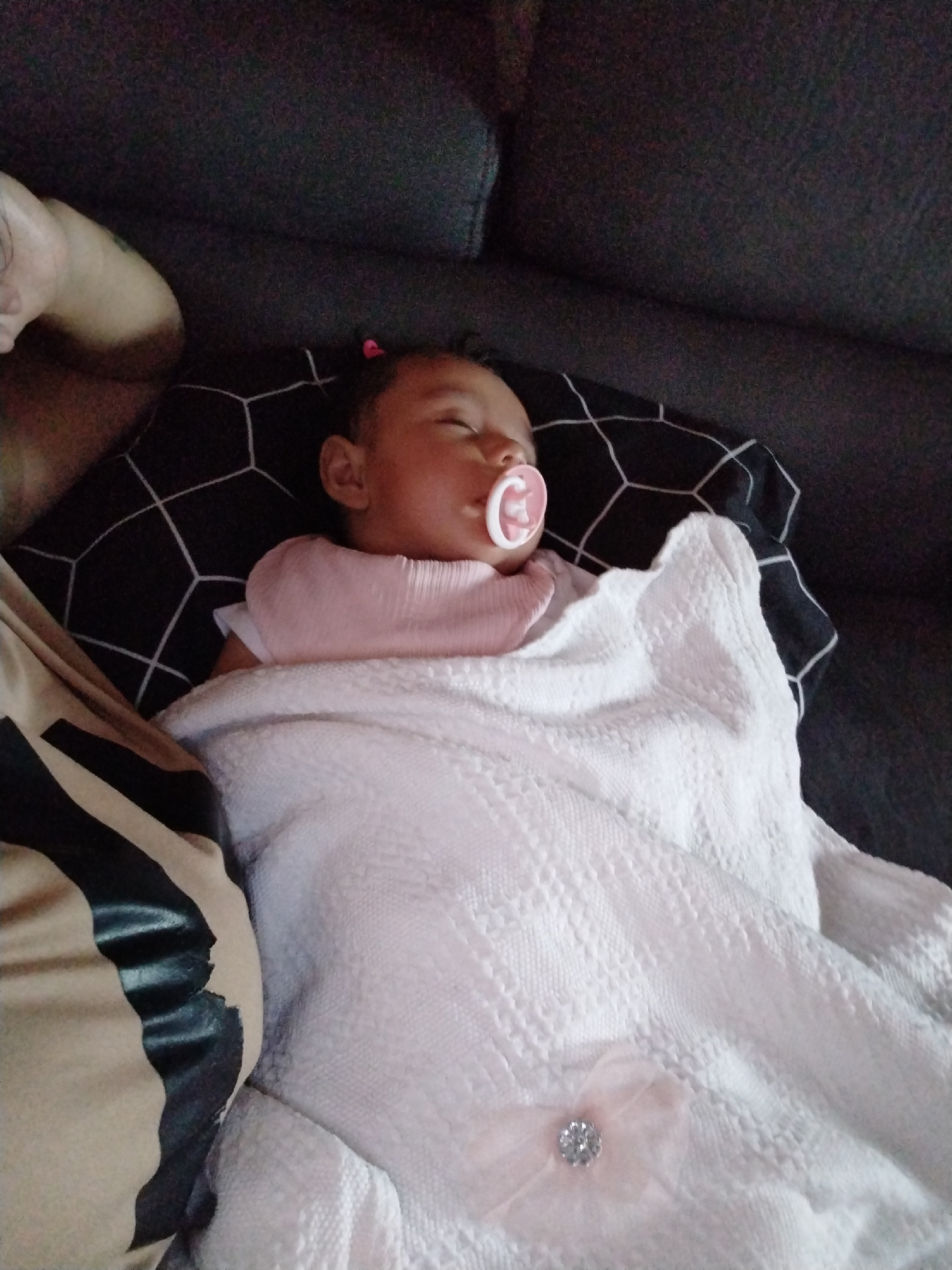 An overhead indoor shot captures a sleeping infant positioned on a dark grey sofa in Unknown, Unknown. The infant, appearing to be a baby girl with closed eyes, wears a pink bib-like top and has a pink pacifier in her mouth. A small pink hair tie or bow is visible on the top of her head. She is partially covered by a white, textured knit blanket adorned with a small pink fabric bow featuring a clear rhinestone center. The baby's head rests on a black pillow with a white geometric hexagonal pattern. To the left, a portion of an adult's tanned arm or shoulder, possibly exhibiting a faint tattoo, is visible, wearing a tan and black patterned garment. The overall lighting is subdued.