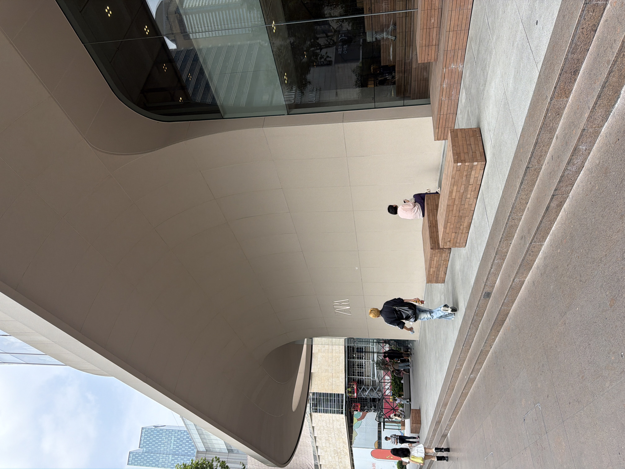 In Qinhuai District, China, an outdoor scene features a large, modern building with a curved, light-brown facade and a glass-fronted entrance. To the right, a woman in a pink top and dark pants sits on a brick bench. A man with blonde hair wearing a black shirt, blue jeans, and a black shoulder bag walks away from the camera towards the building. Further left, a woman in a white dress and black boots walks towards the camera, carrying a yellow bag. Several other people are visible in the background near the building entrance. In the distance, a tall, modern building with a patterned facade can be seen. The ground is paved with stone and concrete, with several steps leading up to a raised walkway with brick benches.