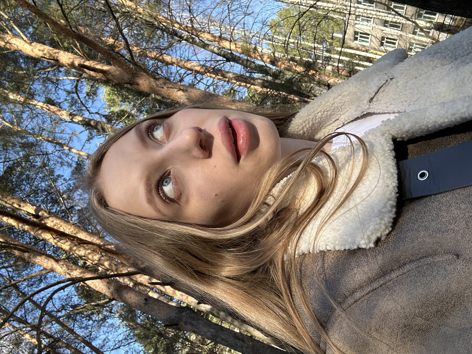 A young woman is photographed outdoors with a low-angle perspective, appearing to look up at the sky. The location is in a wooded area in the city of Chaikovsky, Russia, with pine trees visible in the background along with an urban apartment building. She has fair skin, light blonde hair, and is wearing a brown jacket with a white, fleece-lined collar. The photo is taken in natural light, possibly during the day.