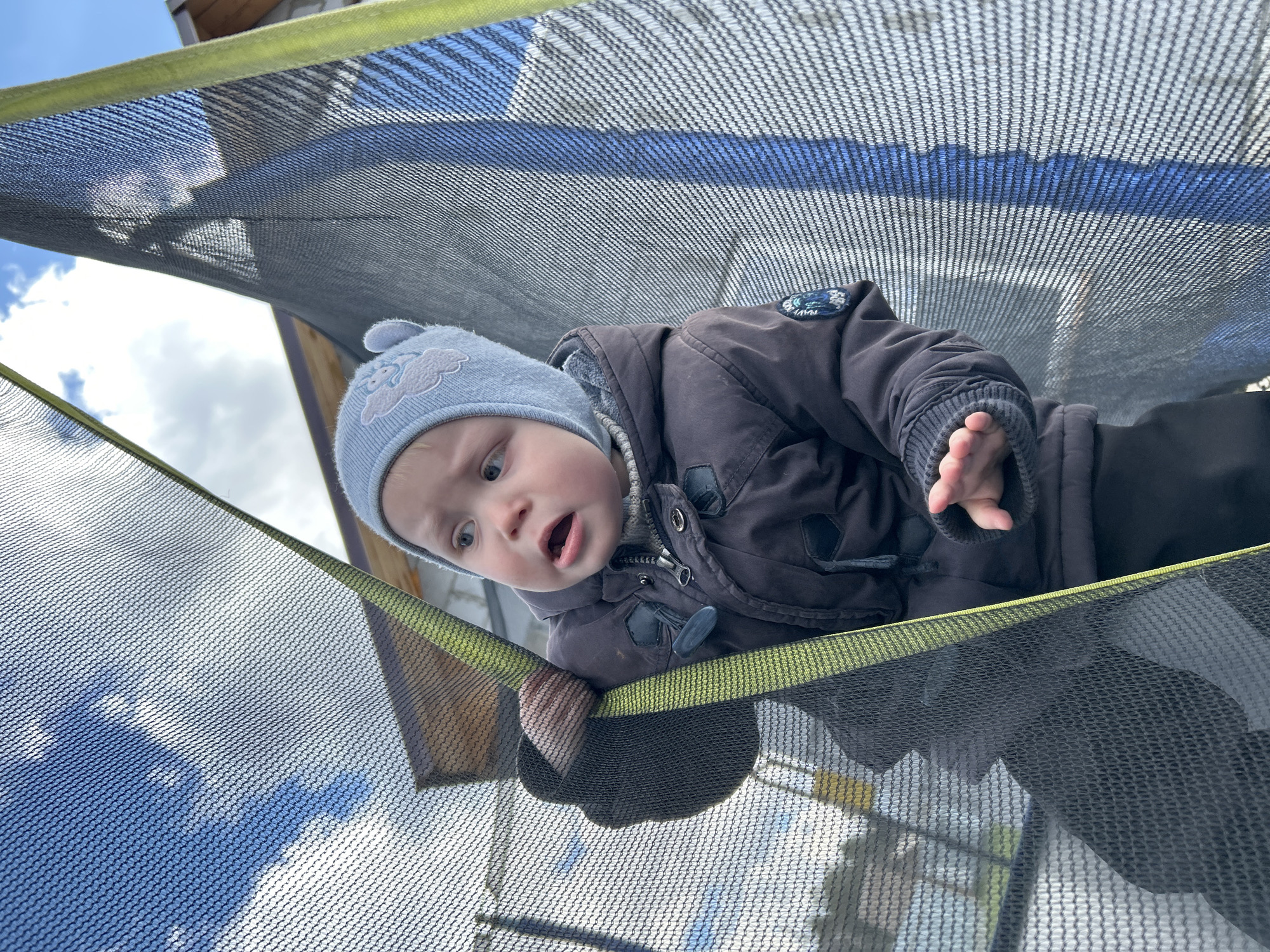 An outdoor, eye-level, low-angle shot captures a young child looking down from within a trampoline's safety net in Chaikovsky, Russia. The child, likely a baby or toddler, is dressed in warm winter attire, including a light blue knitted hat adorned with a white cloud design and a dark brown or grey winter jacket. A small, circular badge, possibly with the word "NAVY" or a similar logo, is visible on the jacket's shoulder. The child's light eyes are wide with a curious or slightly surprised expression, and their mouth is open. One hand is visible, gripping the yellow-green padded edge of the trampoline net. The black mesh of the safety net forms a prominent foreground and midground element, creating a patterned filter over the background. Through the netting, a bright blue sky with scattered white clouds is visible, suggesting a clear daytime setting. Part of a building or wooden structure can be seen in the upper background. The overall impression is of a child observing their surroundings from an elevated, enclosed space.