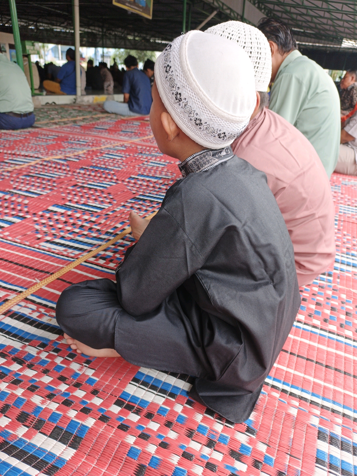 In Manunggal, Indonesia, a group of people, including children and adults, are seen sitting on a woven mat. The mat has a red, white, blue, and black checkered pattern. In the foreground, a young boy wearing a white skullcap and a dark grey robe is seated, facing away from the camera. He is wearing dark grey pants and his bare feet are visible. To his right, an adult wearing a pink shirt is also seated, seen from the back. Further in the background, other individuals are seated, some wearing blue shirts and white skullcaps, and others wearing light green and patterned shirts. The scene appears to be outdoors, under a covered structure with visible supports and roofing. The environment suggests a gathering or event, potentially religious in nature, given the attire and the setting.