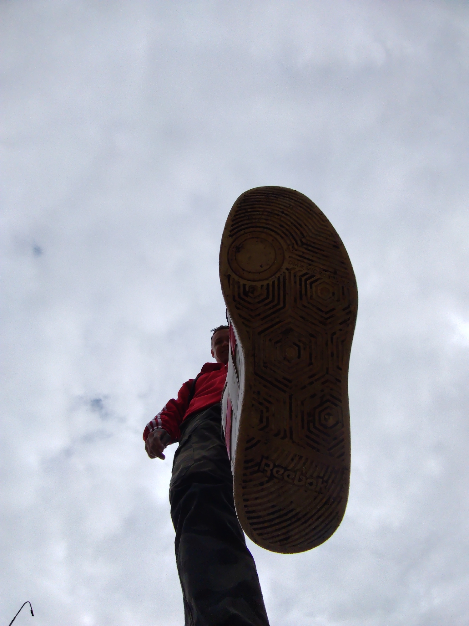 A low-angle shot shows the sole of a Reebok shoe dominating the frame against a cloudy sky. Below the shoe, a person in a red jacket and camouflage pants is visible. The location in Chaikovsky, Russia, is not discernible from the image. The shoe sole exhibits a hexagonal pattern and the brand name "Reebok." The person is looking down.