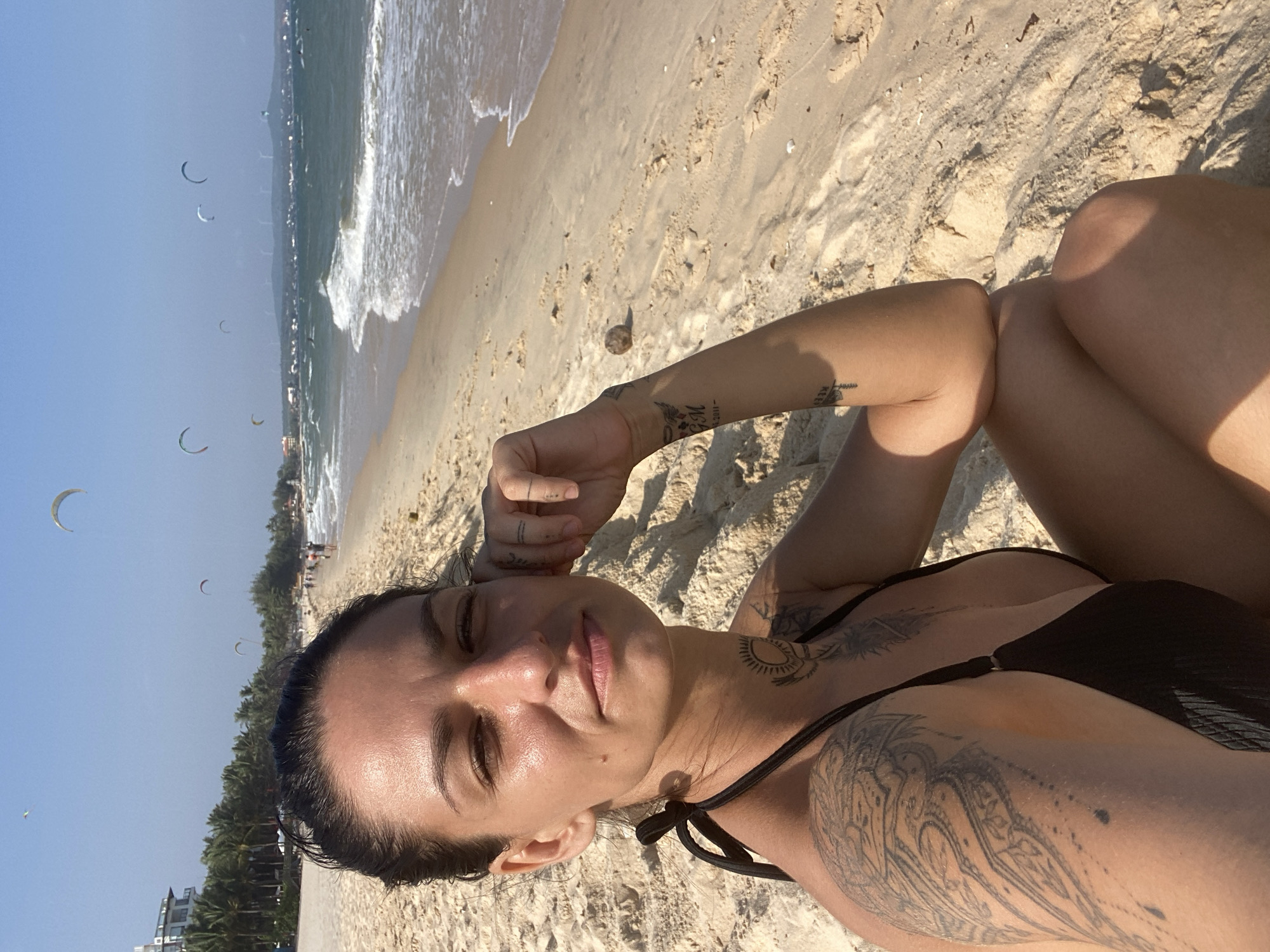The image depicts a woman seated on a sandy beach in Phu Thuy Ward, Vietnam, facing the viewer. She has dark hair pulled back and wears a black bikini top. Visible tattoos include a large, intricate design on her left upper arm/shoulder, a circular sun-like motif on her neck, and a smaller emblem with "G M" and a date "-2011" on her right inner forearm. Her right hand rests near her temple. The beach features golden sand, with gentle ocean waves breaking on the shore to the right. In the background, numerous kites for kitesurfing are active in the clear blue sky above the ocean. A coastline with buildings, including a prominent white multi-story structure and a line of palm trees, extends along the left side. Distant hills or mountains with multiple wind turbines are visible on the horizon, beyond which other people are present on the beach.