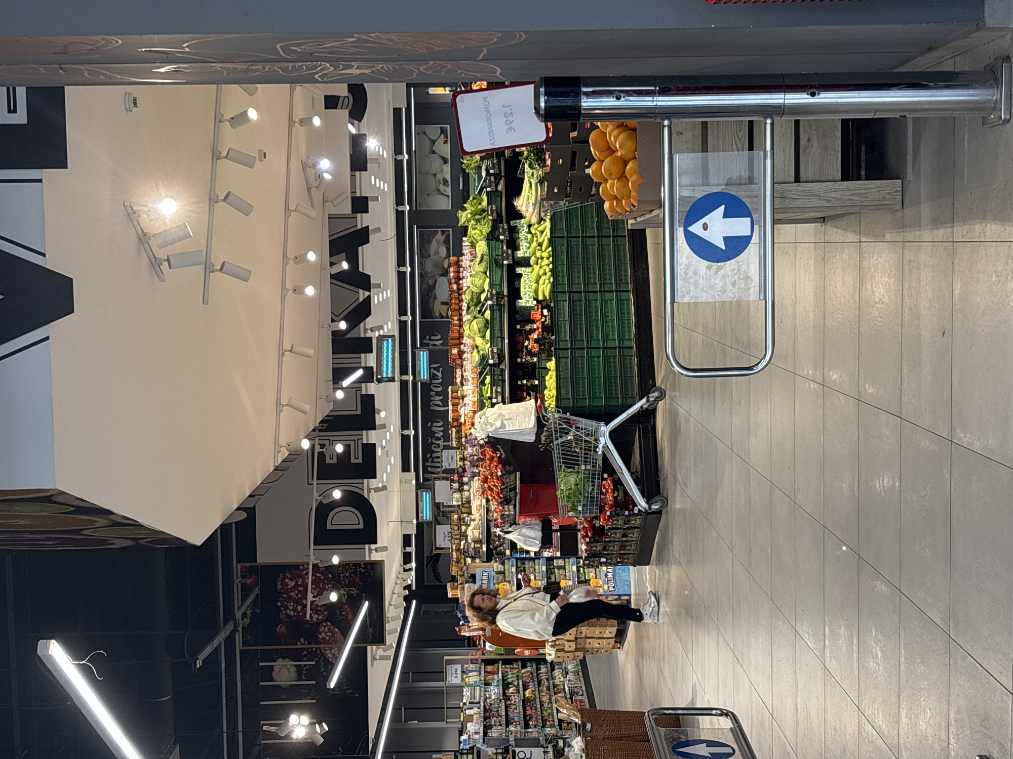 This image captures the interior of a modern supermarket, focusing on the produce and deli sections. The perspective looks down an aisle, with the image rotated 90 degrees clockwise from its original orientation.
In the foreground, on the right side of the main aisle, a chrome barrier features a blue circular sign with a white arrow pointing to the left, indicating customer flow. Beside this barrier, fresh produce displays are visible, including abundant green vegetables in crates and a large pile of oranges. A white rectangular sign with a red border is attached to a fixture, displaying "JABUKE DOMAĆE 3.99€" (Domestic Apples 3.99€).
Further into the store, a woman with brown, curly hair is positioned on the right side of the aisle. She wears a white jacket and dark trousers, holding a smartphone in her hands. A silver shopping cart, containing some leafy greens and other items, is near her, suggesting she is in the process of shopping or browsing. Behind her, shelves are stocked with various products.
The store is brightly illuminated by numerous overhead track lights and long fluorescent fixtures. The left wall features large black letters on a white background, spelling out part of the word "DELIKATESSEN." Additional shelving units hold a variety of products, with text such as "Mliječni proizvodi" (Dairy products), "POLIMAI," and "MARK" visible on labels and packaging, along with a price tag of "2.99€." The floor is tiled in a light grey pattern. The scene depicts a typical shopping environment during an indeterminate time of day, relying on artificial lighting.