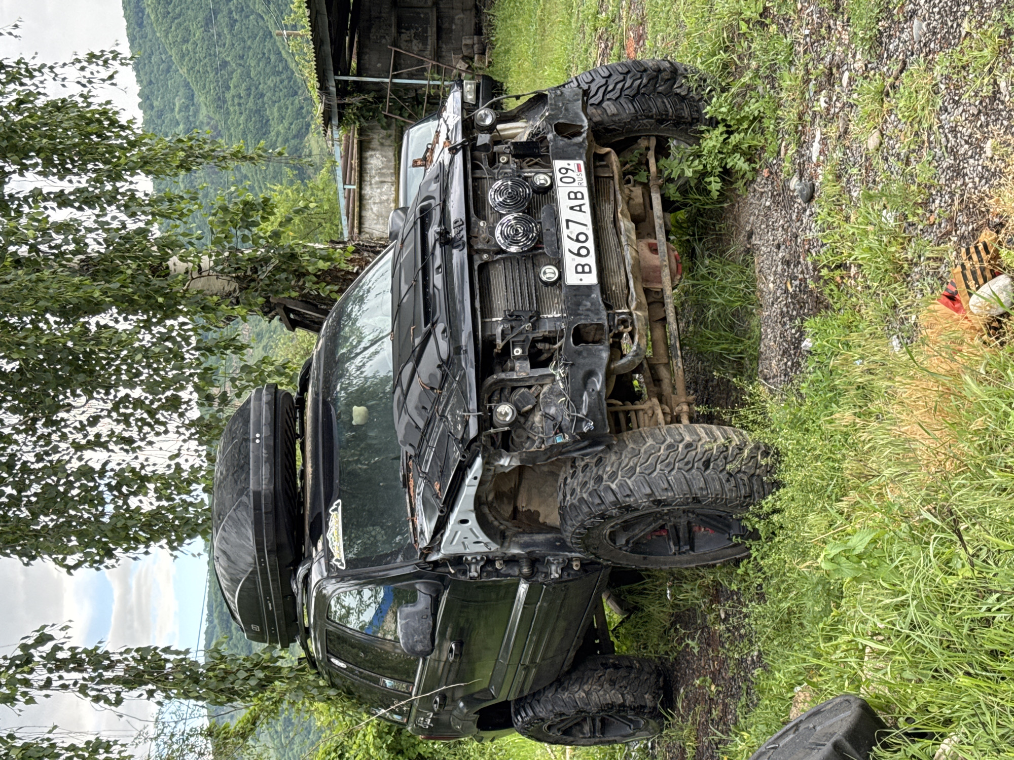 A black, modified Jeep Grand Cherokee with large off-road tires and a roof rack is parked on a gravel and grass area. The front of the vehicle shows significant damage, with the bumper and headlights missing, and the grille exposed. A license plate reading "B667 AB 09 RUS" is visible. The Jeep is situated outdoors, with a tree and lush green foliage in the foreground. In the background, a hillside covered in dense green vegetation and a cloudy sky are visible. A dilapidated shed-like structure is also present in the background. The scene suggests a rural or semi-rural setting, potentially in the city of Karachayevsk, Russia, given the license plate and the mountainous, forested landscape. There are no people or any visible actions or interactions. The image is a static, forensic depiction of the damaged vehicle and its environment.