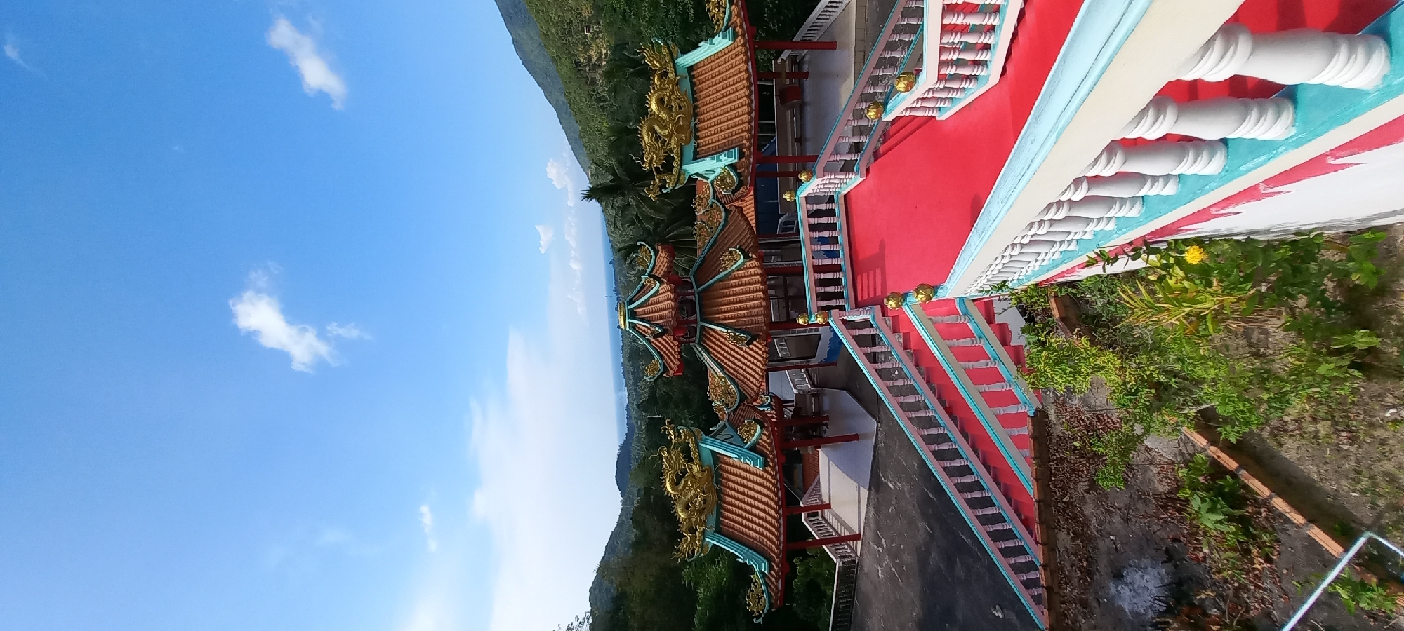 The image, correctly oriented, displays a vibrant, multi-level East Asian-style temple or shrine perched on a lush green hillside under a clear, bright blue sky with a few wispy white clouds.
The central subject is the temple structure, featuring multiple tiered roofs adorned with intricate reddish-brown tiles. Prominent golden dragon figures and other gilded embellishments decorate the roof ridges and eaves, which are painted in striking teal and white. The various levels of the temple are connected by wide, bright red staircases, bordered by white balustrades with teal accents and occasional golden orb-like decorations. Lush green foliage and bushes grow around the base of the structure and along the hillside, with one yellow flower visible near a white balustrade in the foreground.
In the background, rolling green mountains stretch towards a distant, hazy horizon, suggesting a coastal view or a large body of water. The time of day appears to be sunny daytime, likely late morning or early afternoon, with excellent visibility and calm weather. No people or specific activities are depicted. The scene highlights the intricate architectural details of the temple against a natural, expansive backdrop, indicative of its location in Moo 7, Thailand. There is no visible text in the image.