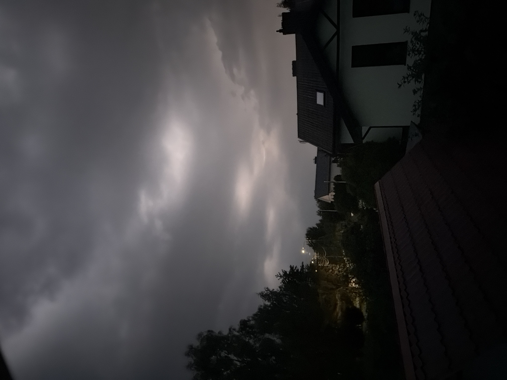 The image depicts a residential area at dusk or during overcast weather, likely in Vilnius, Lithuania. The sky is dominated by dark, heavy clouds with some lighter patches indicating diffused light. In the foreground, the dark, tiled roof of a house is visible. To the right, a white house with dark-trimmed windows and a dark roof is partially seen, with a chimney on top. In the background, other houses and trees are silhouetted against the sky. A street light is illuminated, casting a yellowish glow. The overall atmosphere is somber and muted, suggesting either the approach of a storm or the fading light of day. No people or specific landmarks are identifiable to pinpoint the exact location within Vilnius.