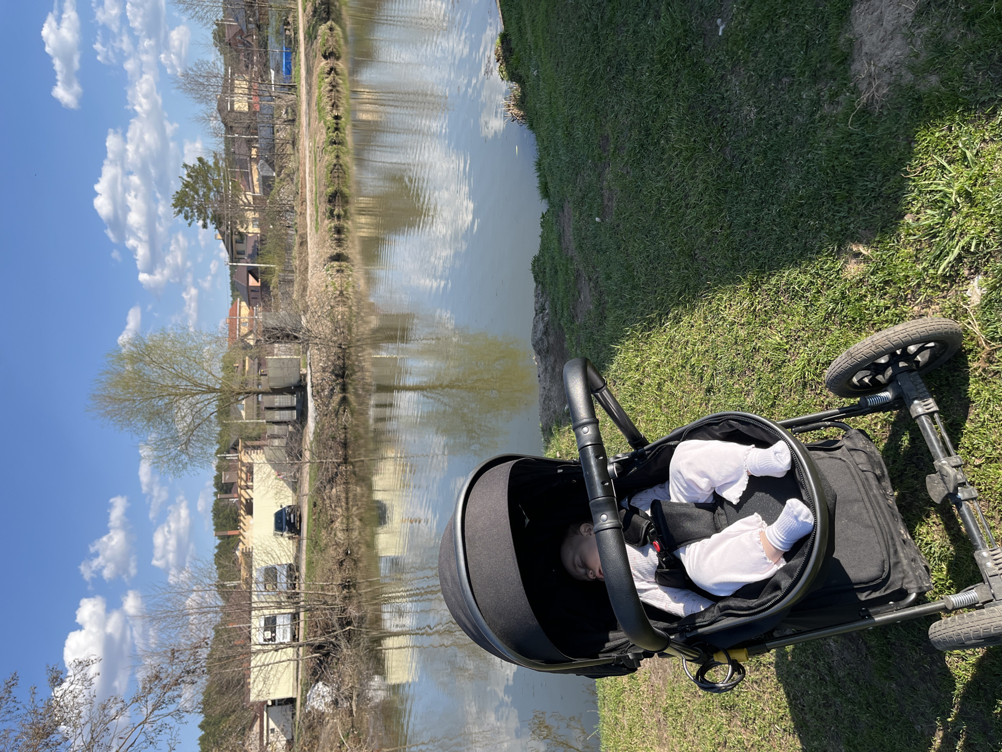 A horizontal outdoor shot shows a baby sleeping in a black stroller on grass next to a body of water. The water is reflecting the sky, trees, and buildings across the way. The background reveals the cityscape of Chaikovsky, Russia, with residential buildings and trees visible along the waterfront. The baby is wearing white clothing and socks. The stroller is angled towards the right. The sky is blue with scattered white clouds. There is green grass on the right side of the scene.