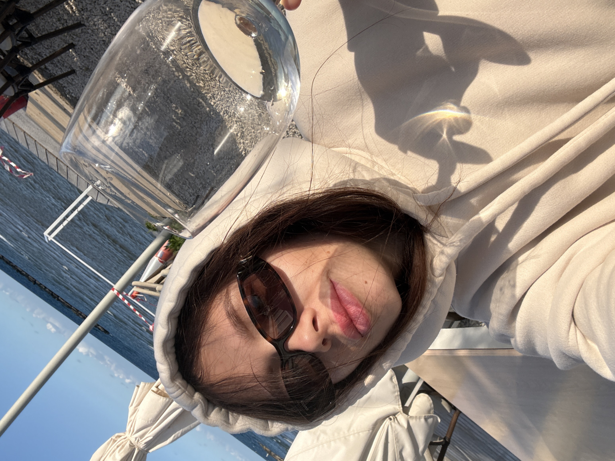 A woman with dark hair, wearing a cream-colored hoodie and dark sunglasses, holds a glass of water in front of her face in an outdoor setting. The background includes a view of the sea, a marina with boats, and elements of outdoor seating, suggesting a restaurant or cafe. The photograph may be taken on the coast of Oroklini, Cyprus, where there are coastal restaurants or cafes.