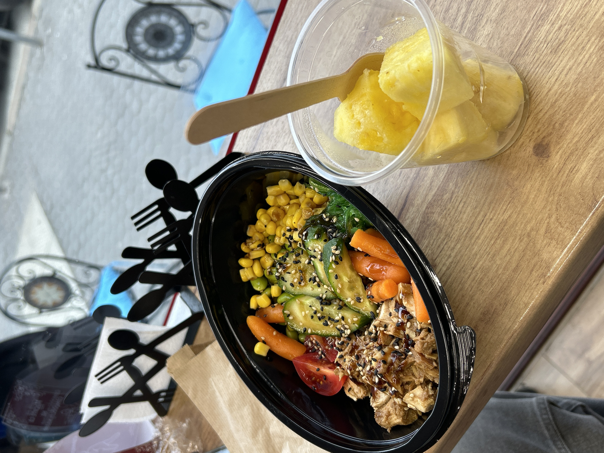 A top-down, slightly tilted shot captures a meal on a wooden surface, potentially a table. To the left, a black plastic container holds a salad comprised of corn kernels, seaweed, sliced zucchini, baby carrots, tomato wedges, and what appears to be shredded chicken or tofu, all sprinkled with black sesame seeds. Beside it, a clear plastic container filled with chunks of pineapple has a wooden spoon resting inside. Nearby is a black decorative item resembling a fork and spoon design. The background contains a window with an ornate iron design, suggesting a cafe or restaurant setting. The location is likely in Sofia, Bulgaria.