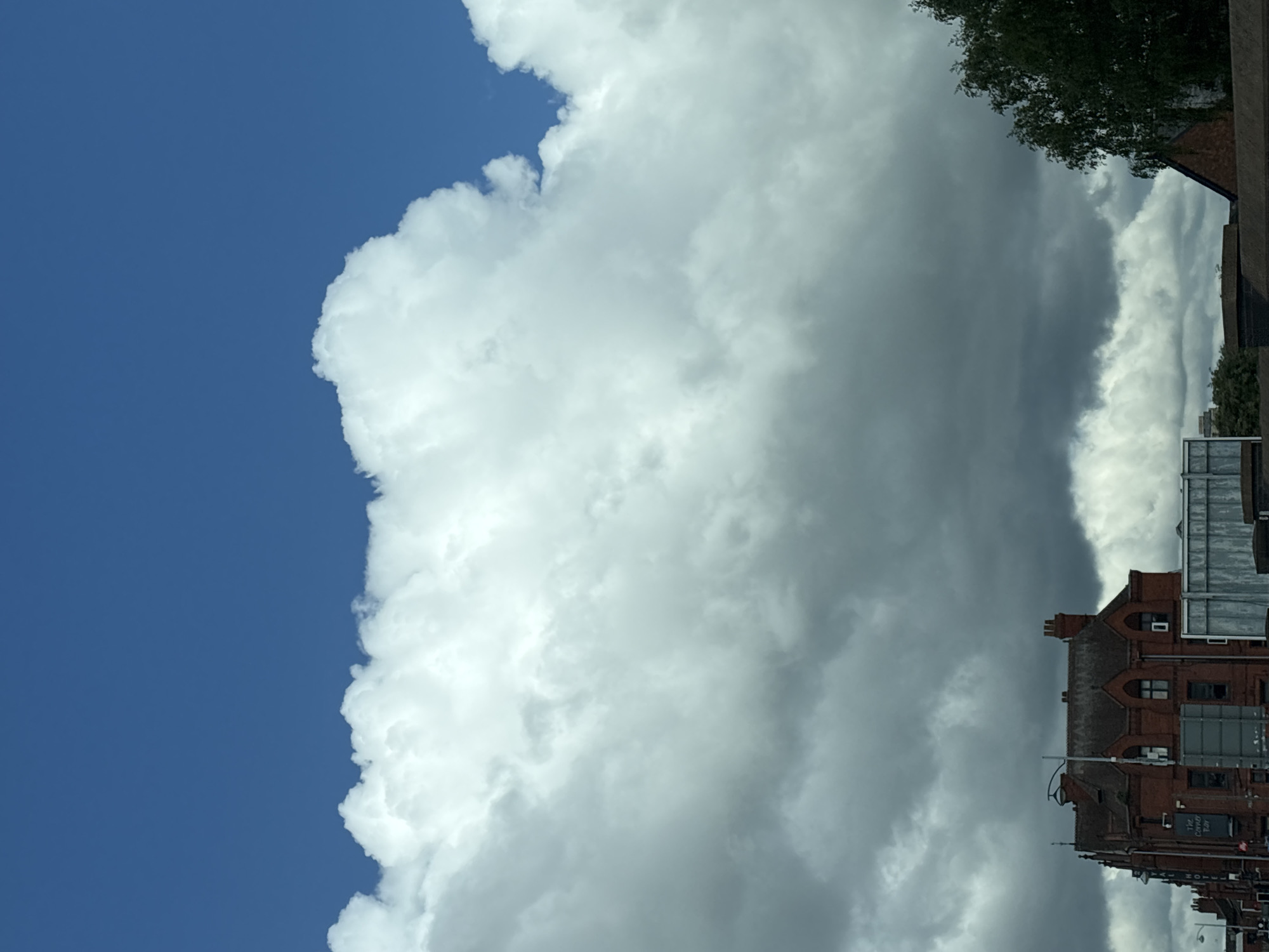 The image shows a large, white cumulus cloud against a blue sky. A red brick building is visible in the lower right corner, appearing to be The Crewe Arms Hotel in Crewe, United Kingdom. Some trees are seen above the building. The image orientation is vertical.