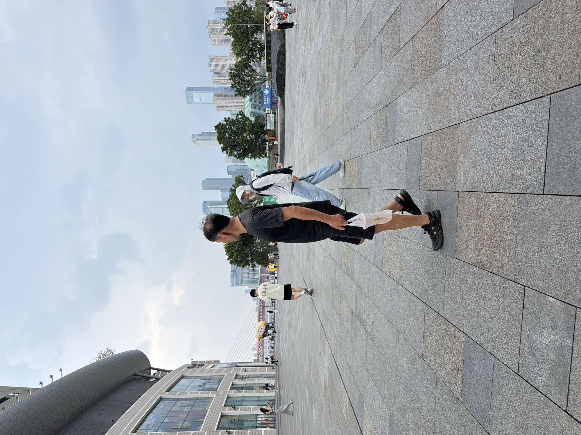 A medium shot captures an outdoor urban scene in Yuzhong District, China, on a cloudy day. The foreground features a man in a black t-shirt and shorts, carrying a white plastic bag, walking on a paved plaza. Behind him, a person in a white hooded jacket, blue jeans, and a backpack walks away from the camera. Further in the background, a crowd of people are visible, some walking, some standing. To the left, a large modern building with glass windows is prominent. Across the plaza, other buildings and trees are scattered, with a cityscape of tall buildings forming the horizon. In the mid-ground, a large inflatable cartoon character is visible, and a bridge spans the water in the distance. The ground is paved with large, rectangular stone tiles.