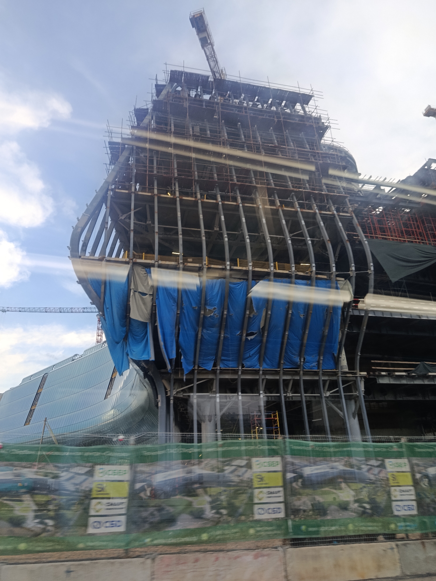 The image shows a large building under construction in Moscow, Russia. The building's skeleton, composed of steel beams, is visible. Blue tarps hang from some sections of the structure. Scaffolding and a construction crane are present. In the background, a curved glass building is partially visible. A construction fence with logos and images is in the foreground. The sky is partly cloudy.