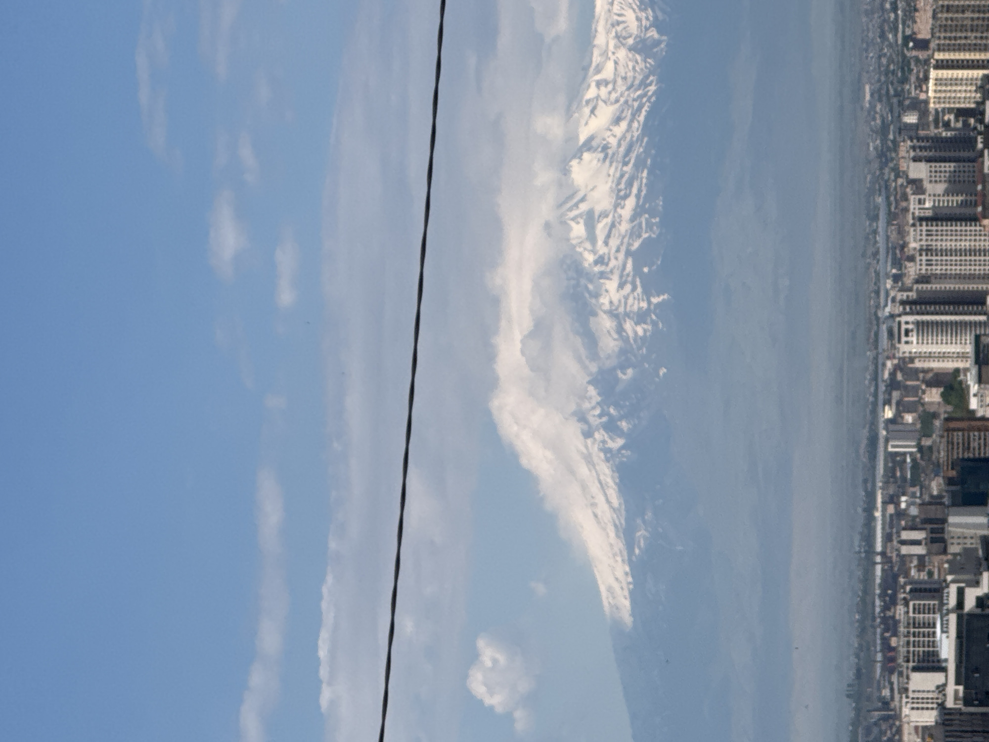 This panoramic landscape image, captured from an elevated vantage point, showcases a vast blue sky, a majestic snow-capped mountain, and a sprawling urban area. The upper and upper-left portions of the frame are filled with a clear, bright blue sky adorned with scattered white, wispy clouds. Dominating the middle-left of the image is a prominent snow-capped mountain range, which, given the context, is strongly identifiable as Mount Ararat, with its rugged, white peaks starkly contrasting against the sky.
To the right of the mountain and extending towards the far right edge of the frame, a dense urban landscape unfolds. This city, identifiable as Yerevan, is characterized by numerous buildings of various heights and architectural styles, creating a complex urban tapestry. A hazy expanse, possibly distant terrain or a body of water, lies between the base of the mountain and the city's edge. A thin, dark cable or power line traverses the upper-left sky diagonally, entering from the top-left and exiting towards the lower-right, adding a foreground element to the expansive view.
The scene is illuminated by bright daylight, suggesting clear weather conditions during the day, likely morning or early afternoon. There are no individuals, specific activities, or visible text present in the image.