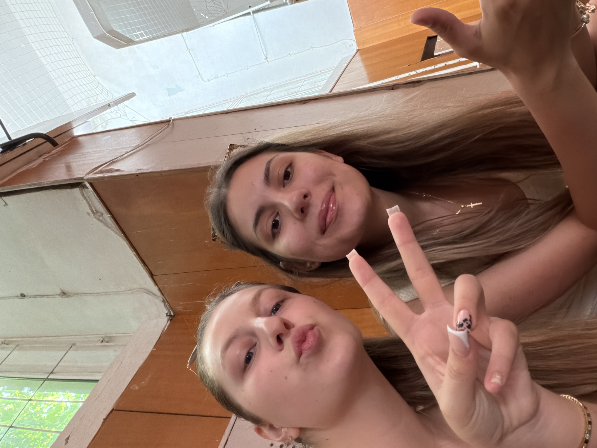 Two young women are taking a selfie. The woman on the left is making a kissing face and holding up two fingers in a peace sign. The woman on the right is smiling and sticking out her tongue slightly, giving a thumbs up with her right hand. They appear to be indoors, with wooden paneling on the walls and a window with greenery visible. The environment suggests a casual setting, possibly a community center or a public space in Burgas, Bulgaria, given the context. The lighting is natural, coming from the window.