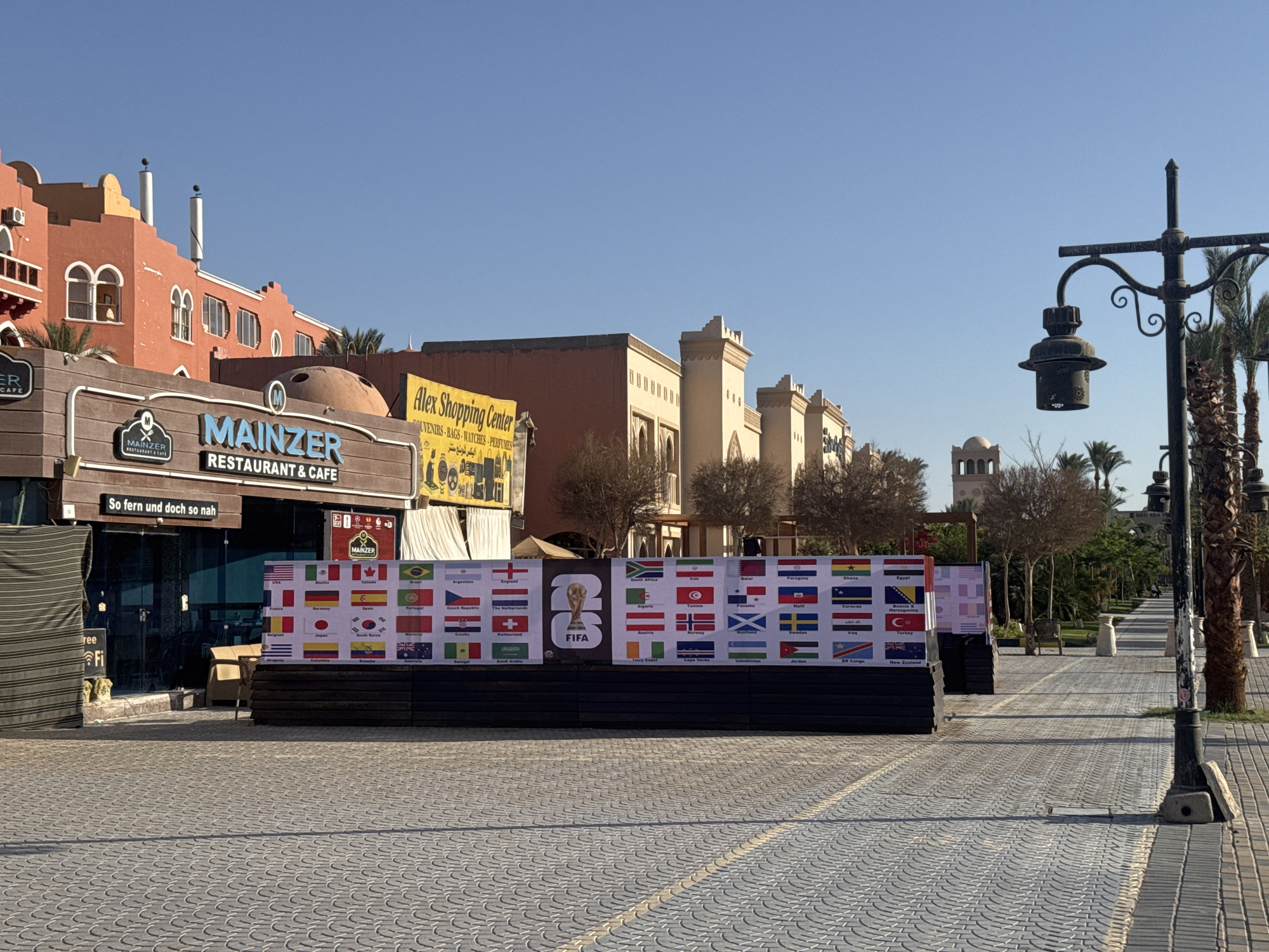 The image shows a street scene in what appears to be a resort area. In the foreground, there is a paved walkway made of interlocking grey pavers. To the right is a vintage-style black lamppost with a hanging lantern, and a row of palm trees. To the left, there is a restaurant named "Mainzer Restaurant & Cafe" with its sign visible, and a closed storefront with a sign reading "Alex Shopping Center" above it.
In the middle ground, a dark barrier is constructed and covered with posters displaying the flags of various countries, as well as the FIFA logo, suggesting the scene is related to the FIFA World Cup. Beyond the barrier, there are more buildings with a mix of architectural styles and colors, including orange and beige. There are also some trees.
The sky is clear and blue. There are no people visible in the immediate foreground. The overall impression is of a tourist area, possibly in a warm climate, setting up for an event. The location is labeled as the city of Unknown, Unknown.