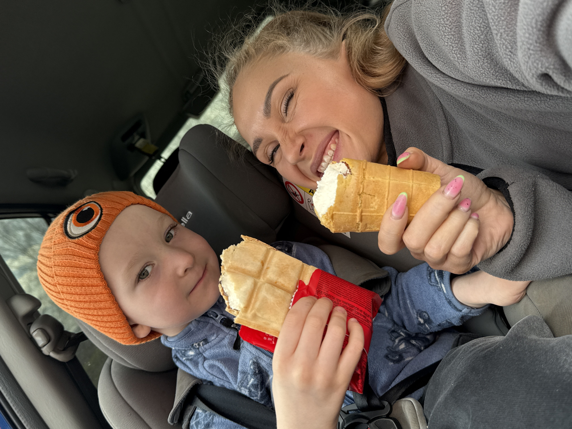 A woman and a child are in a car. The woman is smiling and holding a waffle cone ice cream. The child, wearing an orange hat with a cartoon eye on it, is holding a waffle-shaped ice cream still partially wrapped in a red wrapper. The child is in a car seat. The scene is in the interior of a car. No other details about the city of Krilovo, Russia, are apparent in the image.