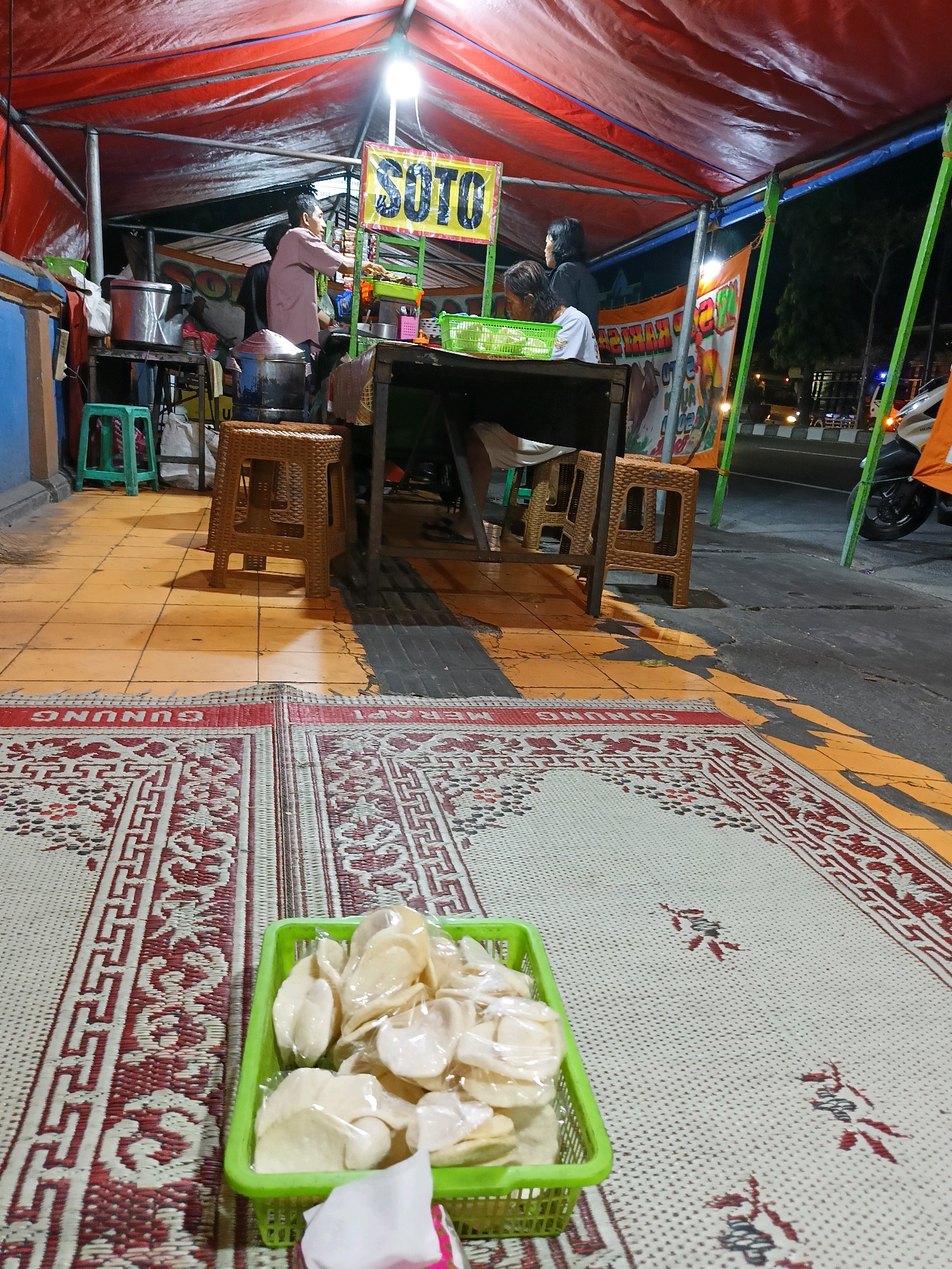 The image captures an outdoor food stall in Kebondalem Kidul, Indonesia, at night. A red tarp shelters the stall, illuminated by a bright lamp hanging overhead. A banner displaying the word "SOTO" in yellow and blue letters is prominently featured. Two women and a man are present at the stall. One woman, wearing a white shirt, is seated at a table. Another woman, with dark hair, stands behind the counter. A man, wearing a light pink shirt, is also behind the counter, appearing to serve customers or prepare food.
In the foreground, a green plastic basket filled with what appear to be crackers or chips in individual plastic wrappers rests on a woven mat. The mat is laid out on an orange-tiled floor, with a section of dark asphalt visible to the right, indicating the stall is situated on a street. A motorcycle is partially visible on the right side of the frame. The environment suggests a casual, street-side dining experience.