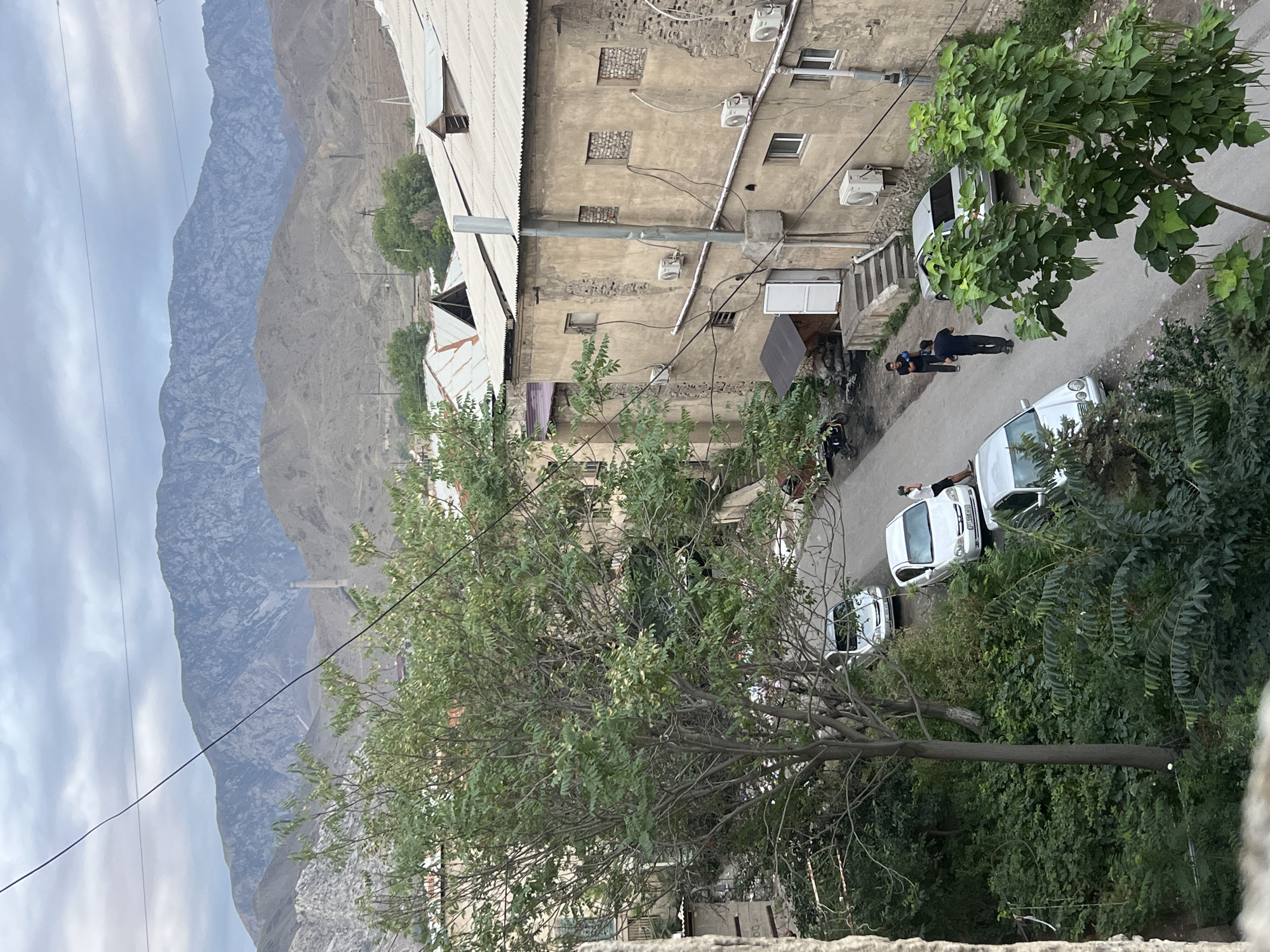 In the city of Kadamjay, Kyrgyzstan, an overhead view reveals a street lined with buildings and vegetation, with a mountainous landscape in the background. The foreground is dominated by a large tree with dense green foliage. The street itself is paved and appears to be in a residential or industrial area. Several cars are parked or driving on the street, including a white sedan and a silver sedan. Two individuals are visible on the street, one standing near a white car and another walking away from the viewer. The buildings lining the street are made of stone and concrete, with weathered facades and corrugated metal roofs. Air conditioning units are mounted on the exterior walls of some buildings. The background features rolling, barren hills leading up to a large, rugged mountain range under a cloudy sky. A tall, slender industrial structure, possibly a smokestack, is visible on the distant hillside. The overall scene depicts a somewhat rugged, urban environment integrated with nature, set against a dramatic mountainous backdrop.
