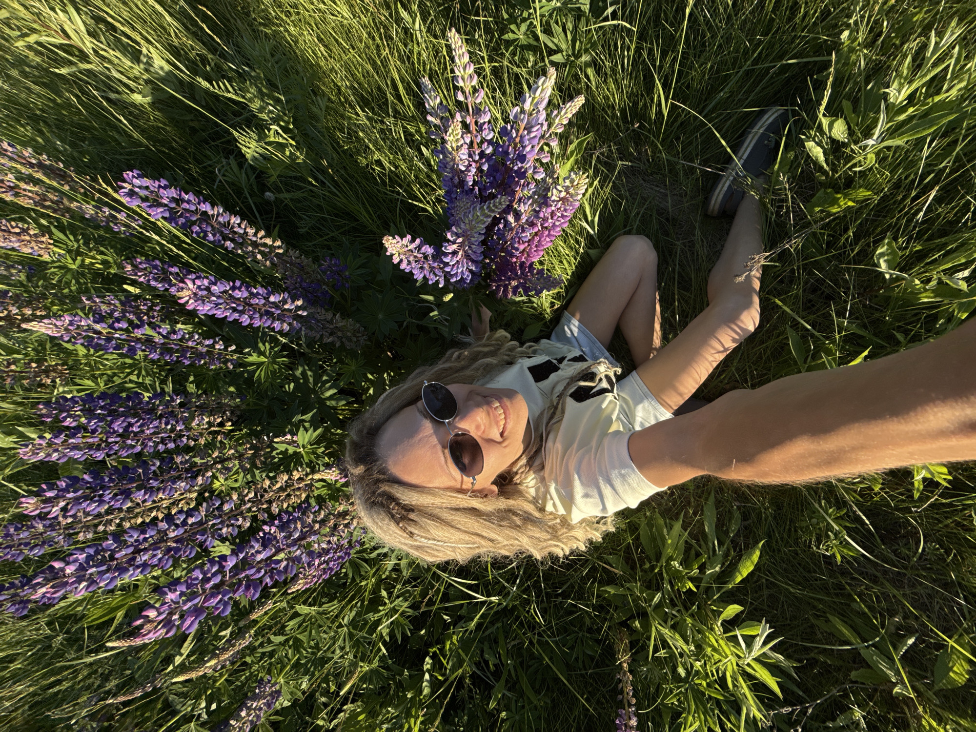 A single adult female is centrally positioned in a high-angle selfie shot, kneeling in a field of tall green grass and blooming purple lupine flowers in Заволжье, Russia. She possesses blonde, wavy hair with braided sections, wears dark oval sunglasses with silver frames, and exhibits a broad smile, revealing her teeth. Her attire includes a light-colored short-sleeved t-shirt and light blue denim shorts, with dark shoes featuring light stripes visible on her right foot. Her right arm is extended, holding the camera to capture the image. A small collection of cut purple lupine flowers is placed on the grass near her left side. The scene is illuminated by direct sunlight, producing defined shadows across the vegetation and highlighting the vibrant colors of the flora.