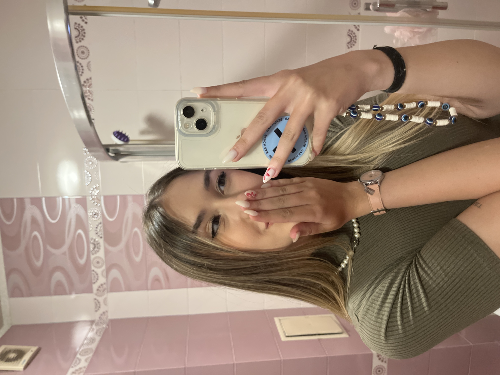 A young woman is captured in a mirror selfie taken in a bathroom in Sofia, Bulgaria. She has long, light brown hair with blonde highlights and is wearing an olive green ribbed top. She holds a smartphone, likely an iPhone, in a clear case with her left hand. Attached to the back of the phone is a light blue circular grip, featuring reversed text that reads "BEACH BABE" multiple times around a stylized 'X'. Her nails are long and light-colored, with a small red heart design visible on the ring finger of her right hand. This hand is positioned to partially cover her mouth, suggesting a playful or blowing-a-kiss pose.
Her accessories include a pearl necklace, a white and blue necklace adorned with "evil eye" beads, a black wristband on her left wrist, and a watch with a pink strap on her right wrist. A reversed tattoo, appearing to be "TXL," is visible on her upper right arm.
The bathroom features tiled walls in shades of pink and white, some adorned with intricate decorative patterns. In the reflection, a metallic towel rack and a small purple hairbrush can be seen. The lighting indicates an indoor setting.