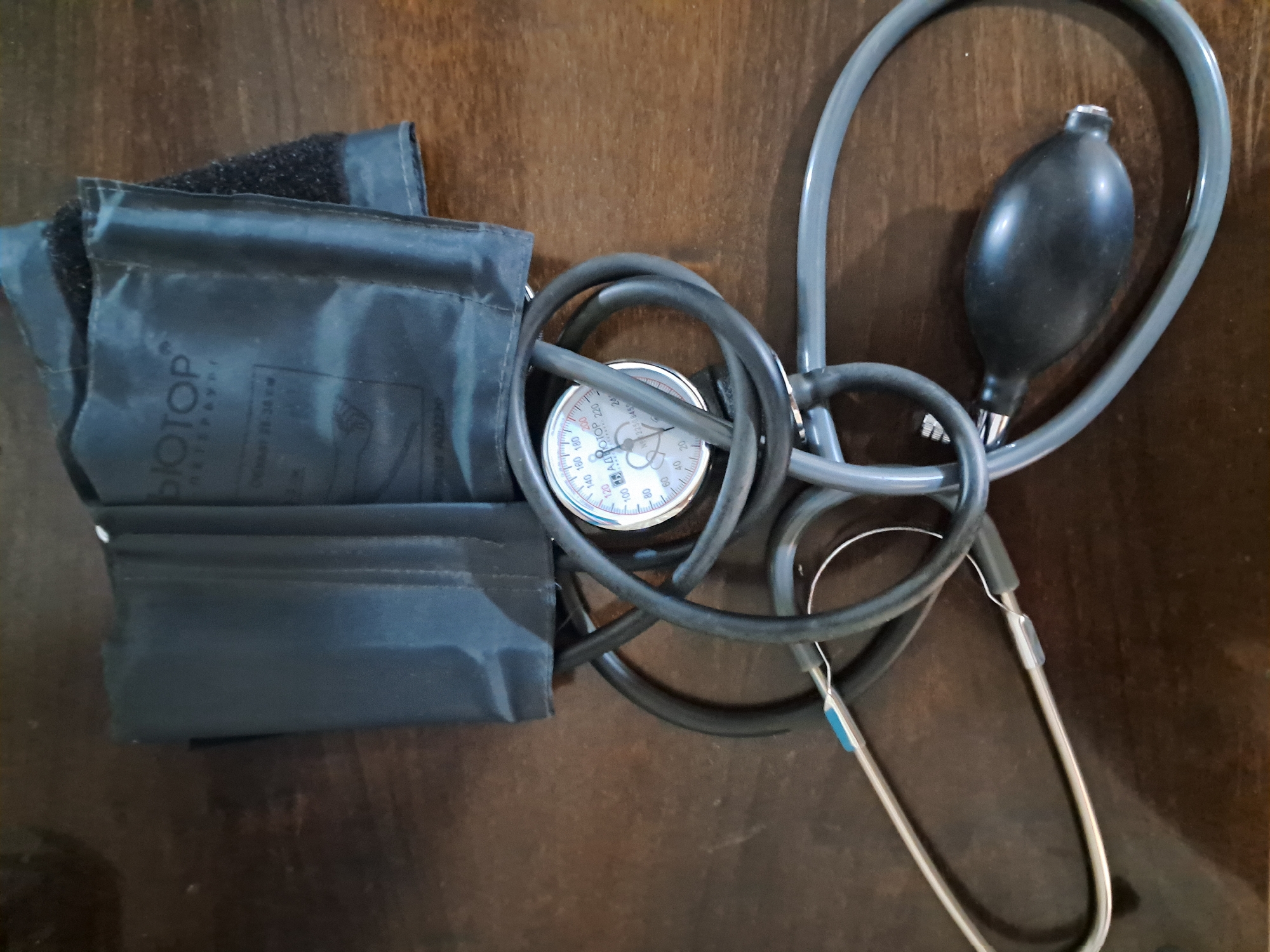 A blood pressure monitor and stethoscope are laid out on a wooden surface. The blood pressure cuff is black and appears to be made of fabric. It has the brand name "BIOTOP" printed on it in white letters. The cuff is rolled up and secured with velcro. The stethoscope is also black and has a grey tube connecting the earpieces to the chestpiece. The chestpiece is circular and silver. A black rubber bulb is attached to the tubing of the blood pressure monitor. The wooden surface appears to be a table or desk. The lighting suggests an indoor setting. The scene is a still life arrangement of medical equipment. The image was taken in Bagish, Uzbekistan.