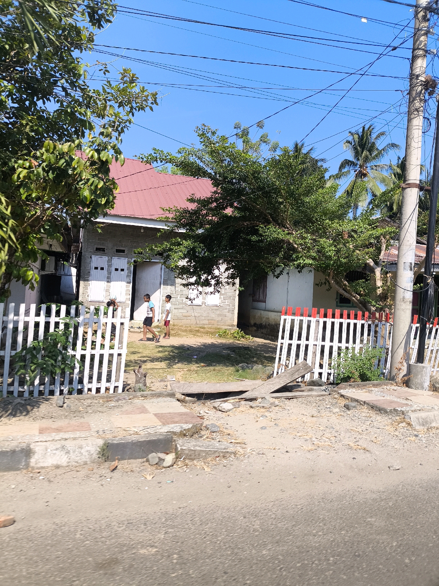 The image depicts a residential scene in Tilamuta, Indonesia, on a clear, sunny day. In the foreground, a rough asphalt road is visible, with a dusty, debris-strewn shoulder. A low concrete curb separates the road from a paved area with checkered tiles, leading to a white picket fence with red accents. Beyond the fence, a modest, single-story house with a red metal roof is situated. The house appears to be constructed of concrete blocks and has multiple windows, some with shutters. Two young boys are standing near the entrance of the house, one wearing a white t-shirt and dark shorts, the other in a white t-shirt and red shorts. They appear to be interacting or simply standing near each other. Trees with lush green foliage surround the house, and palm trees are visible in the background. A utility pole with numerous wires stands to the right of the frame. The overall impression is of a simple, rural residential setting.
