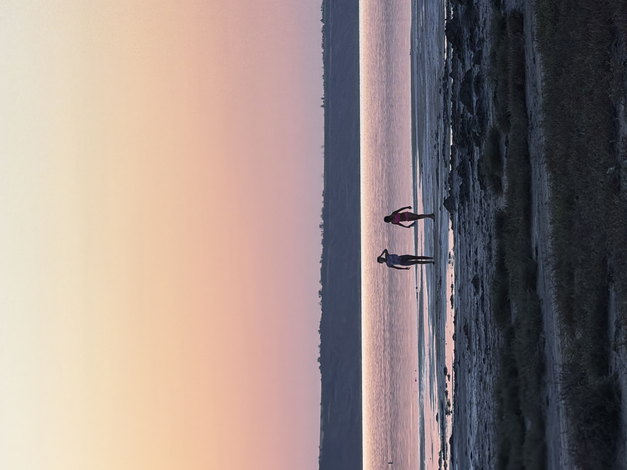 The image depicts a serene twilight scene on the shore of the Kuyalnitsky Estuary in Korsuntsi, Ukraine. Two figures, appearing to be young people, are visible walking along the water's edge. One individual, wearing a dark top and shorts, stands closer to the left side of the frame, with their hand raised to their forehead. The other, in a pink bikini, is to their right, walking with a slight bend in their knees.
The water, reflecting the soft hues of the sunset sky, stretches across the middle ground. The sky transitions from a pale yellow at the top to soft pink and peach tones towards the horizon. A low, dark landmass with silhouetted vegetation forms the distant horizon. The foreground is characterized by a dry, muddy shoreline dotted with small rocks and sparse, dry grass. The overall atmosphere is tranquil and evokes a sense of peaceful solitude.