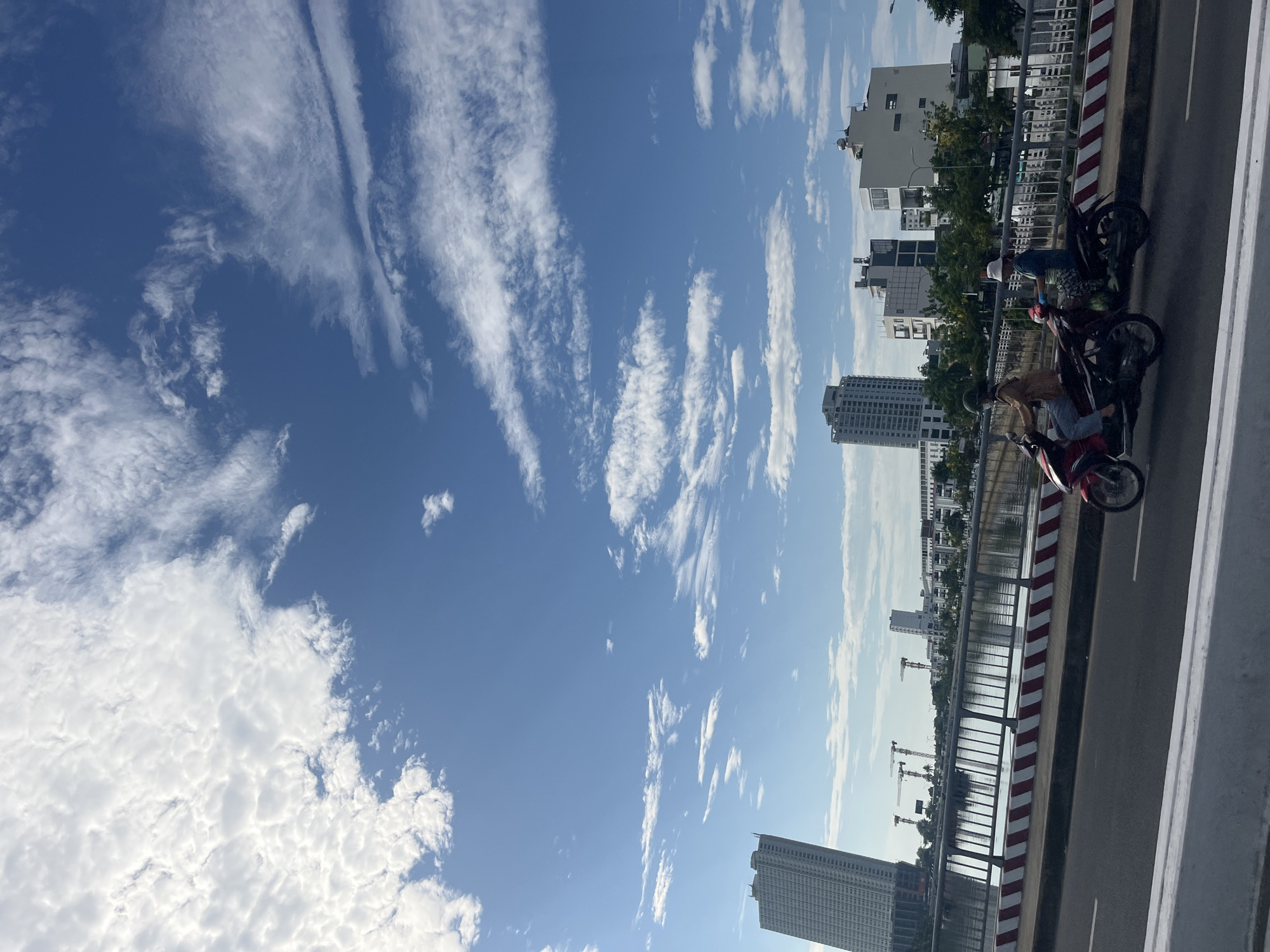 The image presents a vibrant daytime urban scene in Nha Trang Ward, Vietnam, dominated by a vast blue sky and architectural elements. A significant portion of the frame is filled with a bright, clear blue sky, featuring diverse cloud formations. To the left, there are denser, fluffier white clouds, while towards the right and center, long, wispy cirrus clouds stretch across the sky, creating distinct patterns.
Below the sky, the landscape shows a city bridge or overpass running parallel to a body of water, likely a river or canal. The bridge features a metal railing and a distinctive red and white striped barrier marking the edge of the roadway. On the bridge, at least two motorcycles are visible. One person, wearing a beige top and jeans, is seated on a red motorcycle. Another individual, dressed in a blue shirt and a white hat, stands near a dark motorcycle, possibly engaged in a conversation or preparing to ride.
Across the water, a cityscape forms the backdrop, characterized by a mix of buildings. Several modern, multi-story buildings, including some tall structures and various white residential or commercial buildings, line the waterfront. In the mid-ground, multiple construction cranes are visible, indicating ongoing development in the area. The clear weather and strong sunlight suggest it is either morning or late afternoon. No discernible text is visible in the image.