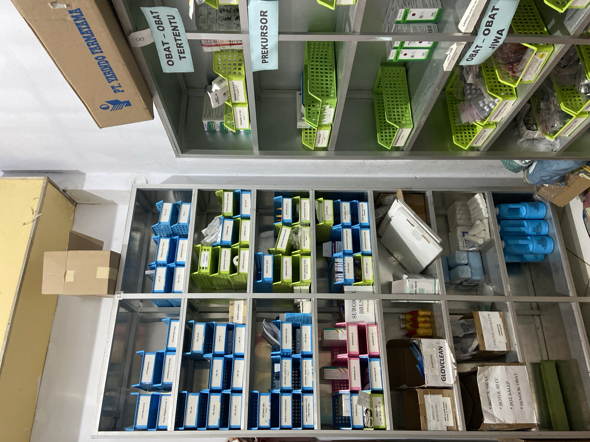The image depicts a meticulously organized storage area, likely a pharmacy or medical supply room, in Margoyoso, Indonesia. Shelves are filled with various medical supplies, neatly categorized and labeled. Prominently displayed are boxes and containers of medications, including bottles labeled "OBAT - OBAT TERTENTU" (Certain Medicines) and "OBAT - OBAT JIWA" (Psychiatric Medicines). There are also trays filled with items such as syringes, medical tubing, and other disposable medical equipment.
Green and blue plastic baskets are used to hold smaller items, and many containers are labeled in what appears to be Indonesian, with terms like "PREKURSOR" (Precursor), "TRAMADOL", "OCTENIC SPRAY", and "GLOVECLEAN". Larger cardboard boxes, some with company logos such as "PT. YARINDO FARMATAMA", are also visible. The lighting suggests an indoor setting, possibly during daylight hours given the overall brightness. The scene conveys a sense of order and preparedness, common in a healthcare or pharmaceutical environment. No people are visible in the image.