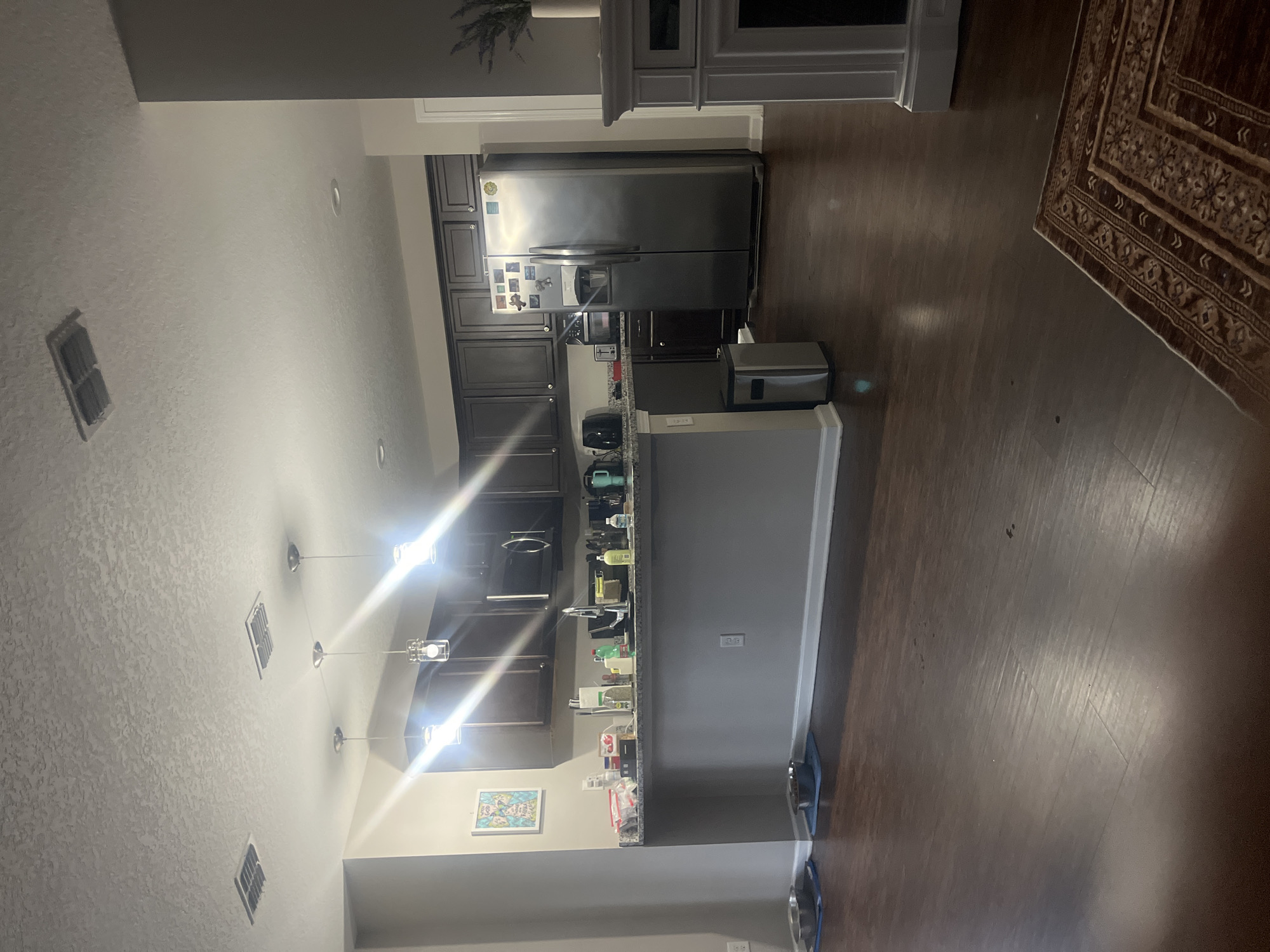 This is a medium-wide shot of a kitchen and living area. The kitchen has dark wood cabinets, a stainless steel refrigerator, and a gray countertop with a sink. There are several light fixtures hanging from the ceiling. In the foreground, the dark wood floor is visible, along with a rug and two dog bowls. A large gray column is on the left side of the image, and a gray fireplace is on the right. The image appears to be taken in a residential setting. The exact location in Jacksonville, United States of America cannot be determined from the image alone.
