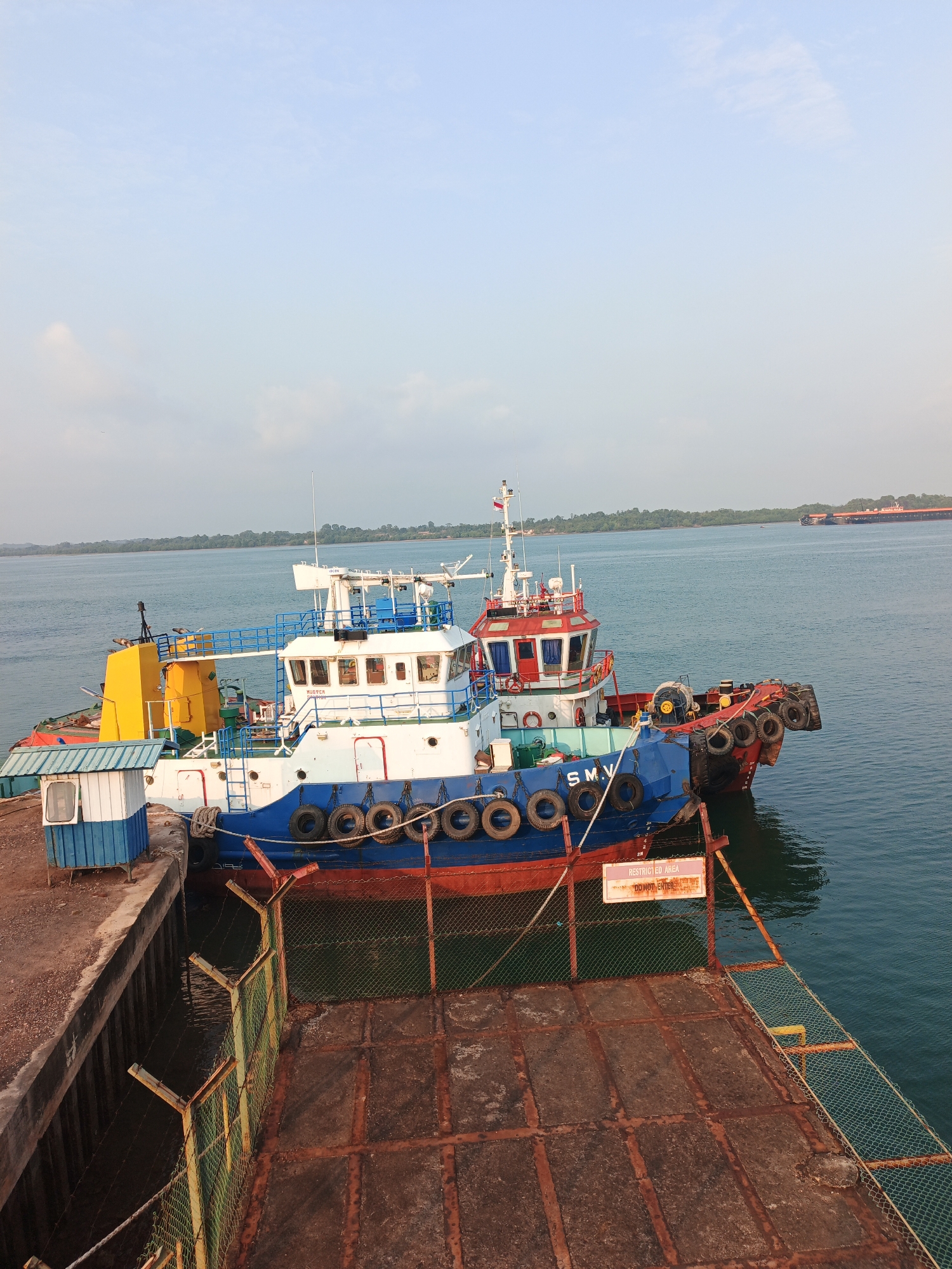 Two tugboats are docked at a pier in Batam, Indonesia. The foreground shows the hull of a blue and red tugboat with multiple tires along its side. A chain-link fence runs along the edge of the pier, with a "RESTRICTED AREA - DO NOT ENTER" sign attached to it. In the background, a white and blue tugboat is tied up next to the first one. The water is calm and a distant freighter can be seen on the horizon. The sky is clear with a few wispy clouds.