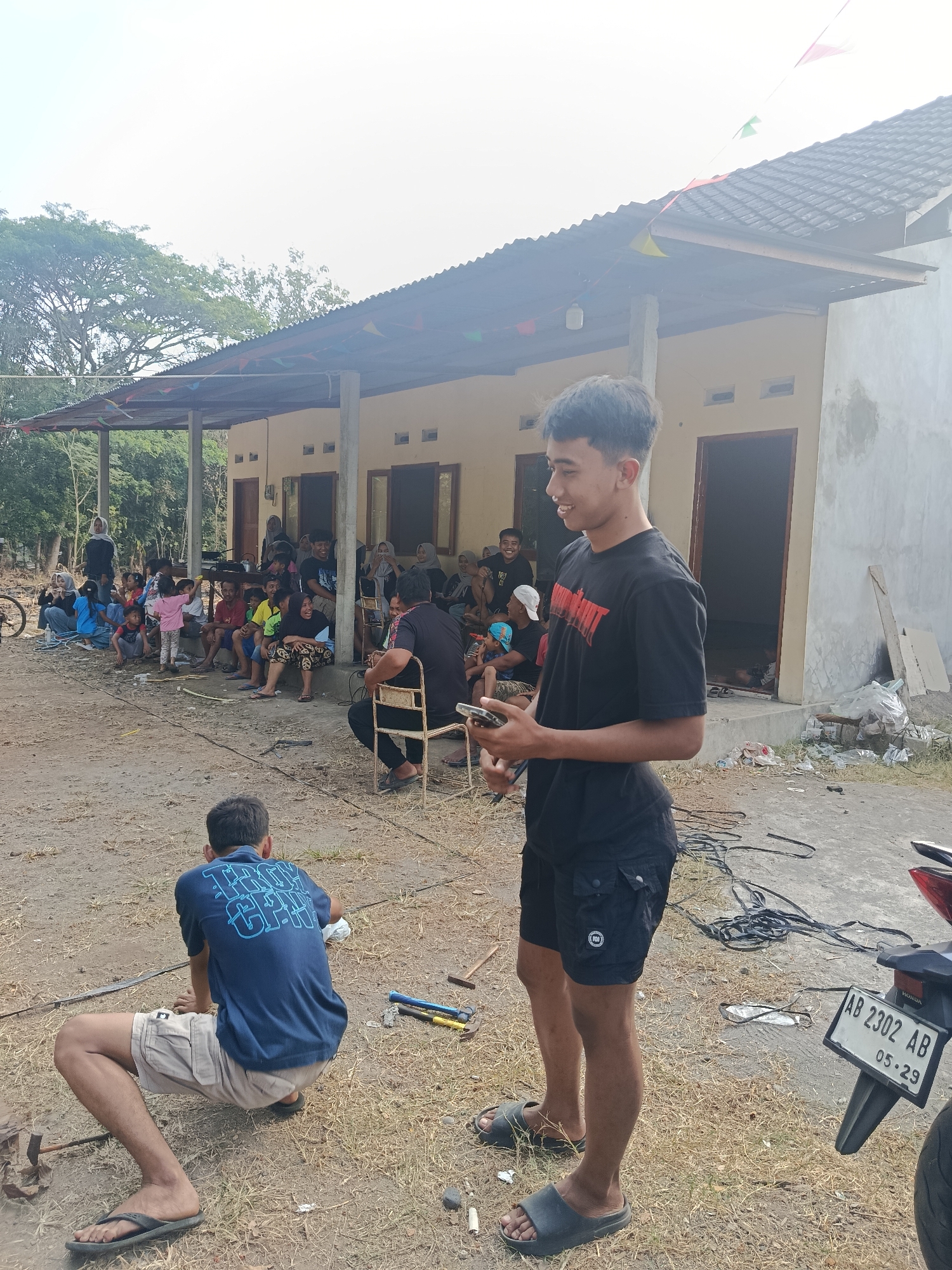 In Sedayu, Indonesia, this outdoor scene depicts a gathering of people in front of a single-story building with a corrugated metal roof supported by concrete pillars. Small triangular flags are strung along the roofline. The building's walls are painted yellow, with some sections showing exposed concrete. Several doorways are visible, some open to the interior.
In the foreground, two young men are present. One is standing, wearing a black t-shirt with a red graphic and black shorts, holding a mobile phone. He is smiling and looking to his left. The other man is crouched down, facing away from the camera, wearing a blue t-shirt and light-colored shorts.
Behind the standing man, a group of people, including men, women, and children, are seated on the ground and on chairs, some appearing to be engaged in conversation or watching something. A motorcycle with a license plate visible is parked to the right. The ground is a mixture of dry earth and sparse vegetation. Various objects are scattered on the ground, including tools, wires, and trash. The overall atmosphere appears casual and communal.