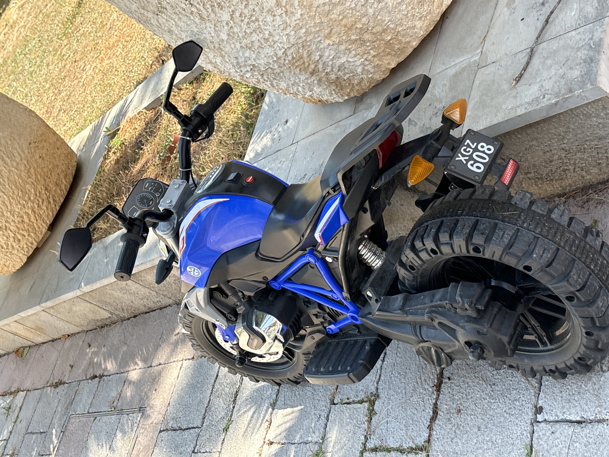 The image shows a blue and white toy motorcycle parked on a stone-tiled walkway in Budva, Montenegro. The motorcycle is oriented vertically in the frame, with its rear wheel closest to the camera. It has a black seat, blue frame, and chrome accents. A license plate with the characters "XGZ 608" is attached to the rear. To the right of the motorcycle, a large, rough-textured boulder is visible, partially obscuring the background. The ground is paved with rectangular grey stone tiles, some of which have grass growing between them. Sunlight casts shadows on the walkway. The scene appears to be outdoors, possibly a public space or a yard.