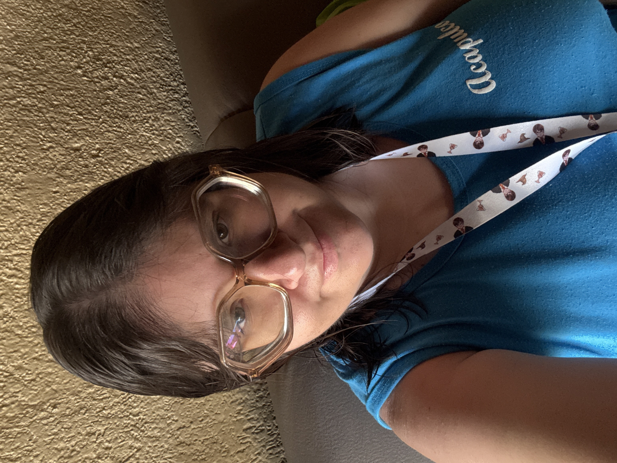 A person, likely female, is captured in a close-up selfie, reclining indoors. They have dark brown hair, which appears slightly damp. Large, clear-framed or light brown-framed glasses are worn, reflecting light from an unseen source. The person looks directly at the camera with a neutral to slightly pleasant expression, revealing their brown eyes, nose, and part of their mouth with light pink lips.
They are wearing a light blue t-shirt with the word "Acapulco" embroidered in white script on the left chest area. The t-shirt fabric shows some pilling or texture. Around their neck is a white lanyard patterned with small, cartoon-style images of Harry Potter, sorting hats, and golden snitches.
The background consists of a textured, golden-brown surface, possibly a stucco wall or heavily textured wallpaper, behind their head. A dark, possibly leather or fabric, armrest or cushion is visible behind their back and shoulder. The lighting suggests an indoor setting. The "Acapulco" shirt and the location context "Tres de Mayo, Mexico" suggest a connection to Mexico.