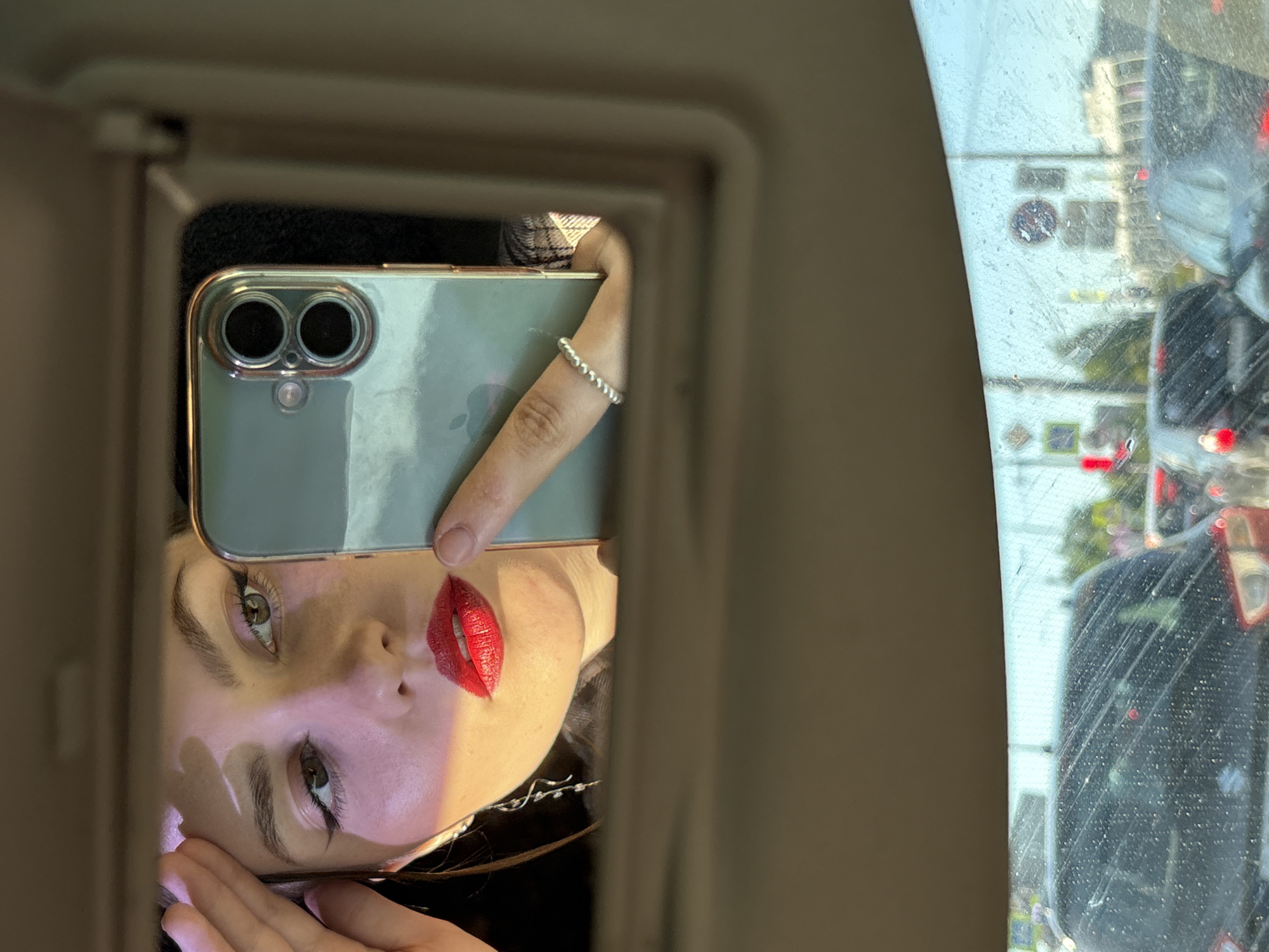 A young woman with light skin, dark hair, winged eyeliner, and red lipstick is visible in a reflection from a car's visor mirror, positioned in the upper portion of the frame. She is using her right hand, adorned with a silver beaded ring, to hold a light green iPhone (likely an iPhone 11 or 12, identifiable by its dual camera system and the Apple logo faintly visible) in a clear case with gold trim. Her left hand rests on her forehead, creating a distinct shadow. A long, silver, dangling earring is visible on her left ear. She appears to be taking a selfie.
The lower portion of the image shows the view through a dirty car windshield, indicating a stationary or slow-moving vehicle. Outside, an urban street scene with heavy traffic is depicted under a bright sky. Multiple dark-colored vehicles with illuminated red taillights are visible. Road infrastructure includes poles and overhead wires. Distinct road signs, consistent with those found in Russia, are discernible: a blue square sign with a white arrow pointing forward (indicating a mandatory direction), a red circular sign with a white horizontal bar (a "no entry" sign), and a partially visible blue square sign with a white 'P' (parking). Red traffic lights are also visible. The environment, including the road signs and general urban landscape, is consistent with a city like Saint Petersburg, Russia.
