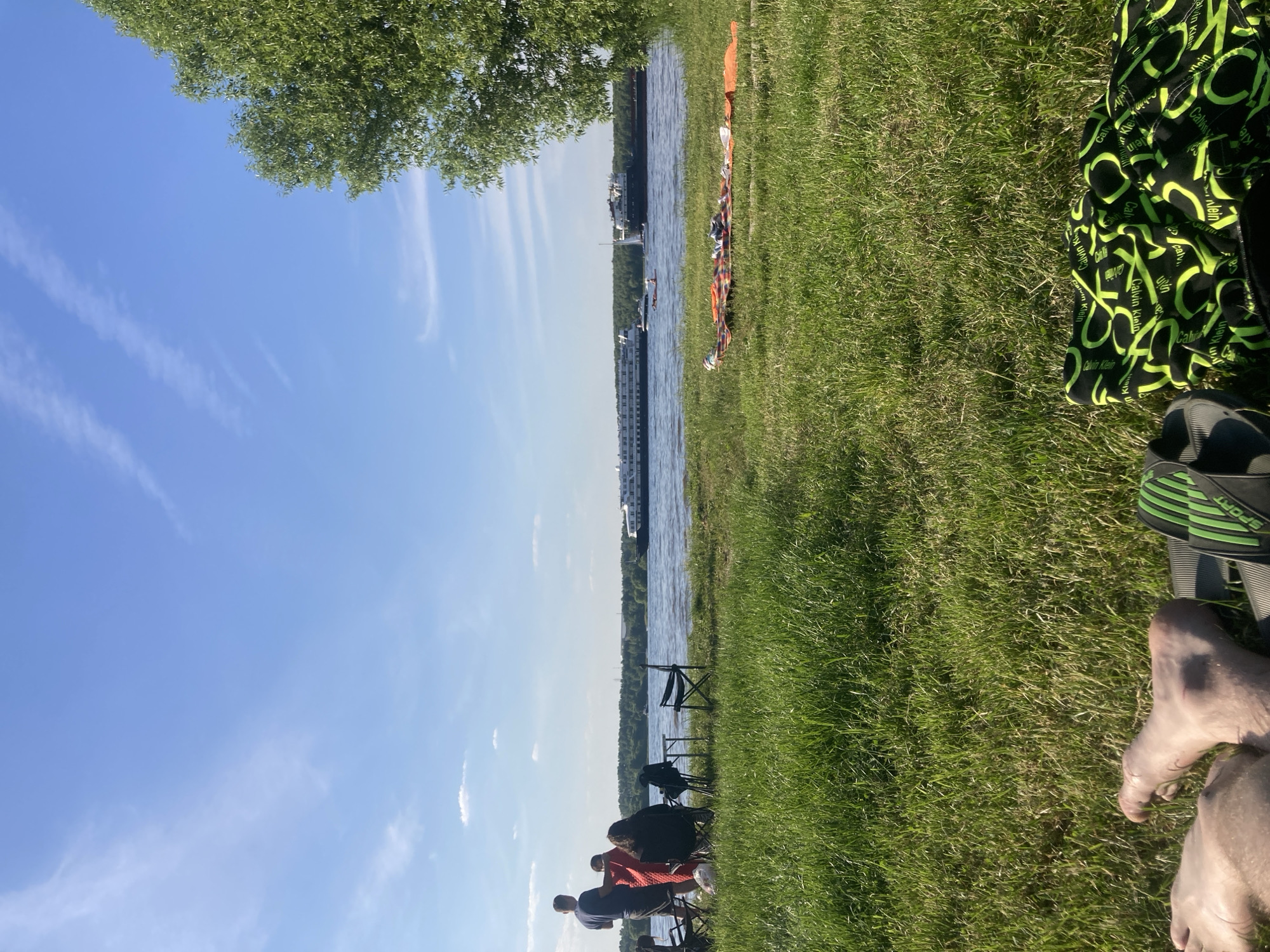 This image captures an outdoor scene along a wide body of water in Kapustino, Russia, under a clear blue sky with sparse, wispy clouds. The sunlight indicates a bright day, likely late afternoon based on shadow definition.
In the immediate foreground, to the left, a person's bare feet are visible, resting on a dark surface, adjacent to a pair of black and green "SPORT" branded flip-flops. To the right, a black fabric item patterned with repeating neon green "Calvin Klein" text is partially visible on the grass.
The midground features lush green grass extending to the water's edge. On the left, a group of four individuals is gathered. One person, seen from the back, stands wearing a dark t-shirt and shorts. Another person is partially obscured, draped in a red/orange blanket. Two other individuals are seated in dark folding chairs, also viewed from behind, positioned near a small, dark portable table. On the right side of the midground, a portion of a leafy green tree is present. Closer to the water on the right, a multi-colored blanket or towel, including an orange section, is spread out on the grass with objects resting upon it.
The background displays a broad body of water, likely a river, with several vessels. Two large, multi-deck passenger ships are prominent; one is primarily white and situated towards the left, while a darker ship is positioned towards the right. Between these larger vessels, a white sailboat with a visible mast and a smaller, low-profile boat containing at least one occupant are present. The far bank is defined by a dense treeline, suggesting a forested area, with faint outlines of distant man-made structures visible above the trees.