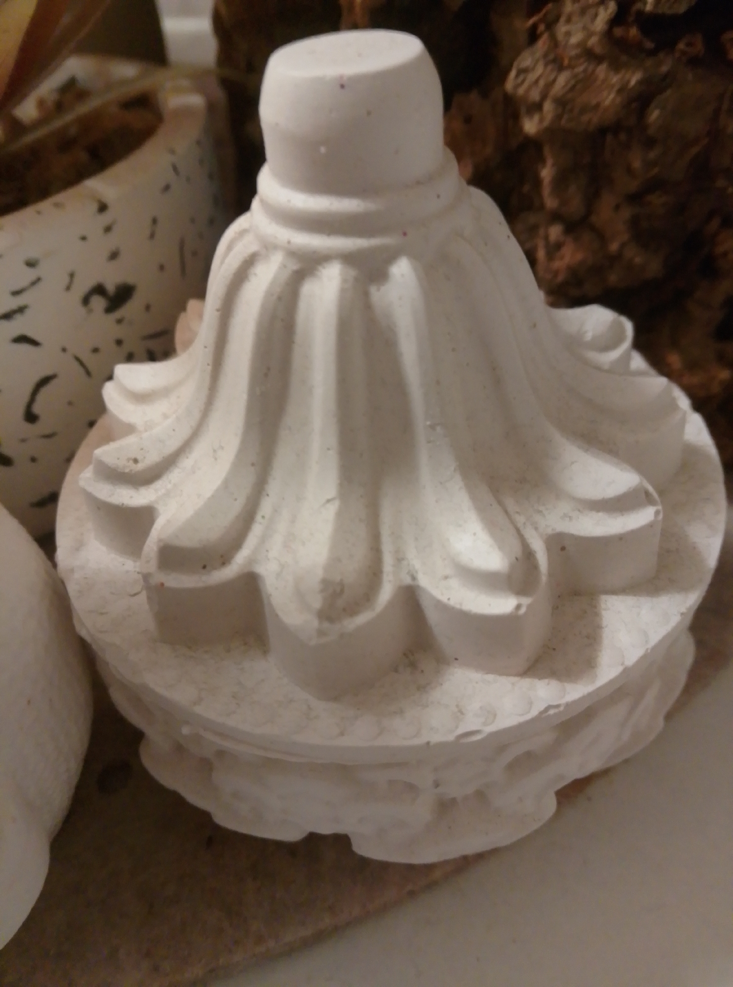 A white, sculpted object, possibly a decorative finial or base, is the primary subject of the image. It exhibits fluted details on its upper portion and a more textured, layered base. The object is situated on a surface that appears to be cork or a similar natural material, with hints of a speckled white pot or planter to the left and a rough, bark-like texture in the background. The lighting is dim, casting soft shadows and highlighting the fine dust or grit covering the white object. There is no indication of the city of Wedel, Germany, within the image itself.
