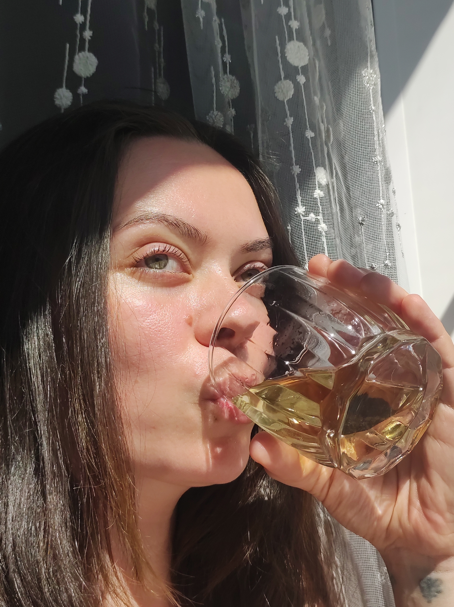 A woman with light skin, dark brown hair, and green eyes is drinking from a faceted glass of a yellow-tinted beverage. She has a small tattoo on her left wrist. Sunlight illuminates her face and hand. She is positioned in front of a window covered with a white, patterned sheer curtain. The location is undetermined and cannot be identified as Фоки, Russia from the image provided.