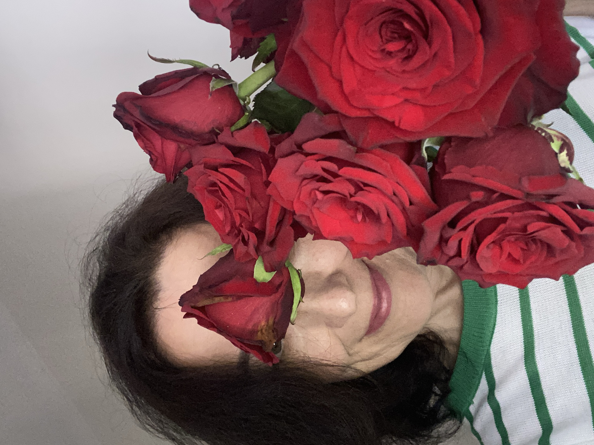 A close-up portrait captures an adult woman posing with a vibrant bouquet of red roses, which are held close to her face, largely obscuring her eyes. The woman has dark hair and light skin, with her nose, mouth, and chin visible. Her lips are slightly parted, hinting at a gentle expression, and appear to have a reddish tint. She is wearing a white top with distinct vertical green stripes. The roses are a deep red hue, with some showing minor signs of wilting or browning on their outer petals. Green stems and leaves are visible among the clustered blossoms. The background is a plain, light-colored surface, suggesting an indoor setting. There are no specific visual cues within the image to indicate a location, but the context provided for this media file is Alanya, Turkey. No text is visible in the image.