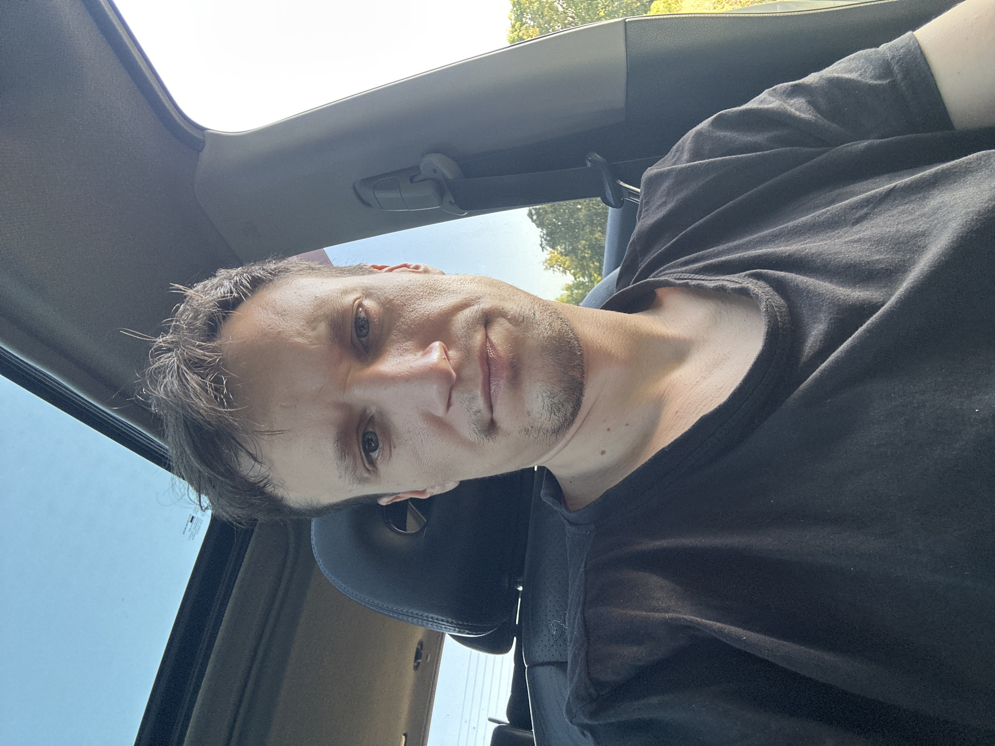 A medium-shot, waist-up, selfie-style photograph shows a light-skinned male with short, messy brown hair and stubble. He is wearing a dark grey t-shirt and is seated in the driver's seat of a vehicle. The vehicle's sunroof is open, revealing a clear blue sky. The headrest of the passenger seat is visible behind him. The background shows a slightly blurred natural environment with green trees and foliage, suggesting an outdoor, possibly rural or suburban, setting. The lighting indicates it is daytime, likely late afternoon due to the warm glow. The image does not contain any specific landmarks or clues to pinpoint a location within Makiv, Ukraine.