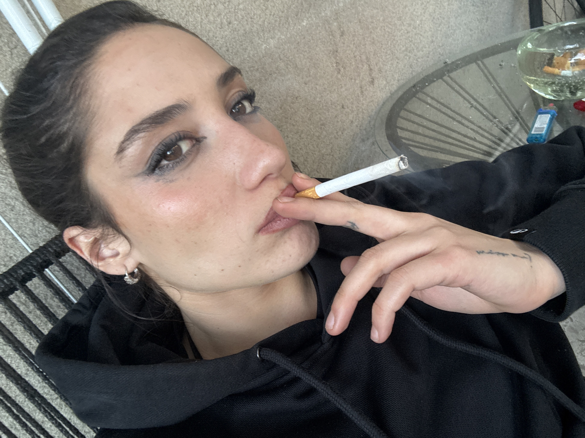 A young woman is photographed from a low angle, holding a lit cigarette to her lips. She has dark hair pulled back, dark eyes, and wears a black hoodie. Her left ear has a silver hoop earring, and a small tattoo is visible on the top of her left hand. A glass-topped table is visible in the background, with a round glass container on it. The container appears to contain cigarette butts and liquid. A lighter and a red object are also on the table. The setting appears to be indoors, possibly on a patio or balcony, as a woven chair is partially visible in the lower left. There is nothing to indicate the specific location is in Buenos Aires, Argentina.