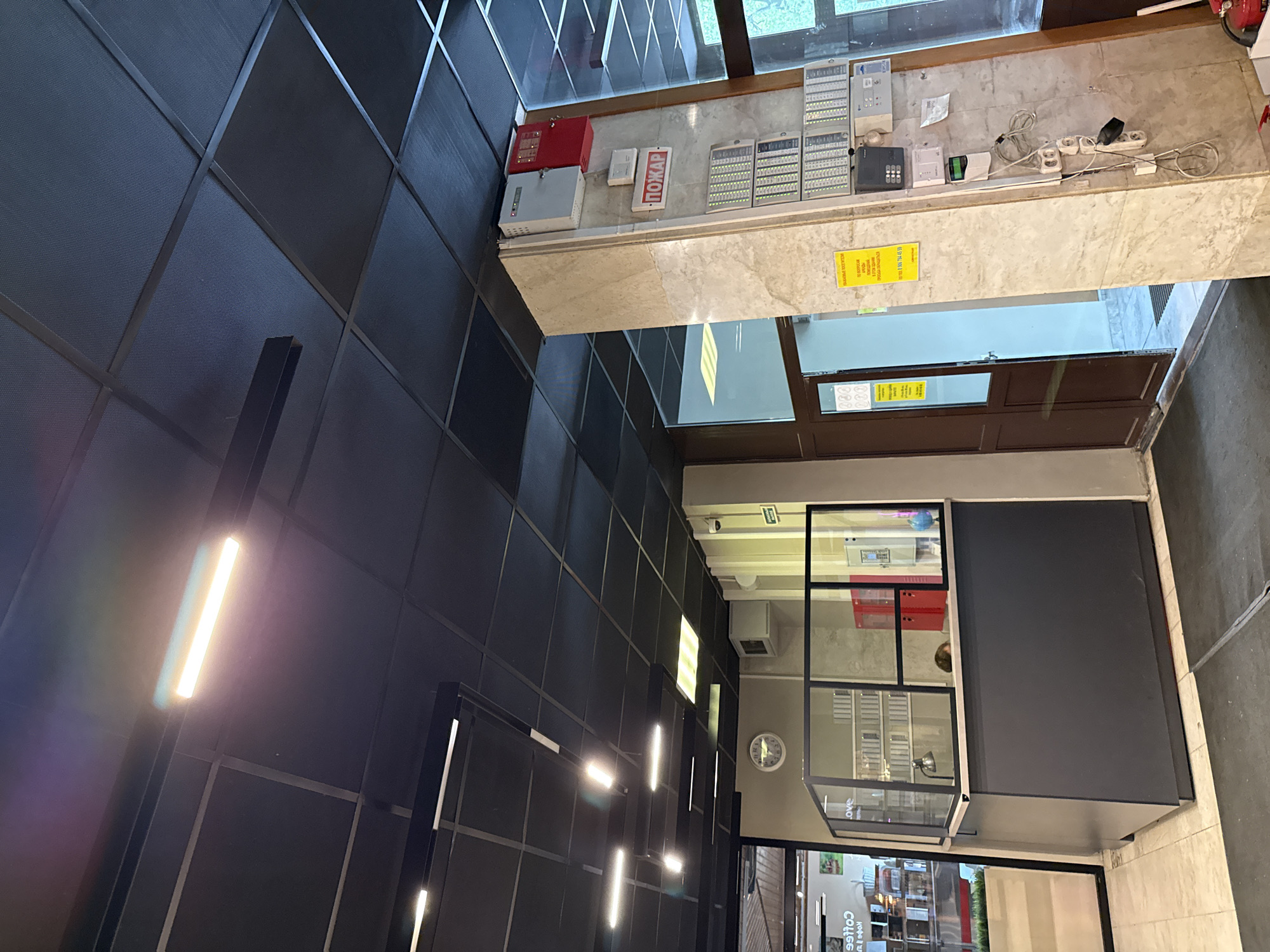 This appears to be an indoor shot, possibly a commercial space. The dominant feature is a grid-like ceiling composed of dark panels with metal framing. Linear light fixtures are integrated within the ceiling structure. High on the right side of the frame, there are security systems, likely related to fire and safety. There is a door and some windows. In the distance, there is a coffee shop, with the word "coffee" printed backwards on the window.
Based on the word "Пожар" (which means fire in Russian) on one of the security items, and a phone number with the +7 country code, this is likely a location within Russia. Considering the modern design, it is plausible that this image was taken in Moscow, Russia, a major urban center.