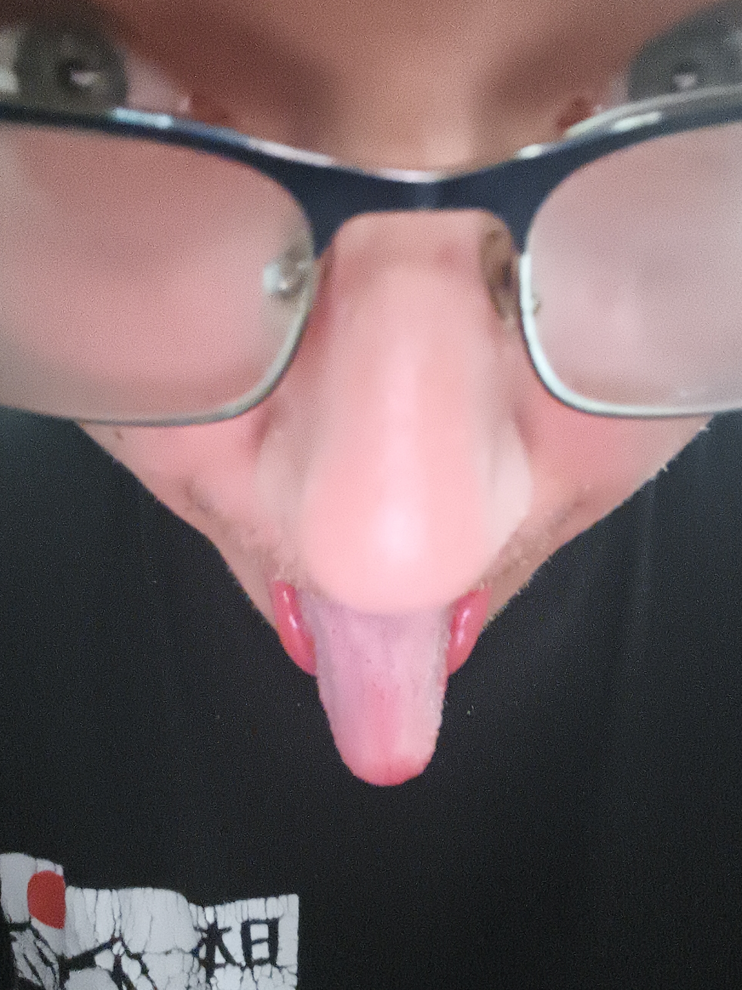 A close-up perspective photo reveals a person wearing eyeglasses, with the image primarily focused on the lower portion of their face, including the nose and mouth. The person is sticking out their tongue. The subject is wearing a black t-shirt with a design featuring a red circle and text written in Japanese or Chinese characters. No details suggest the location is Nowy Tomyśl, Poland.